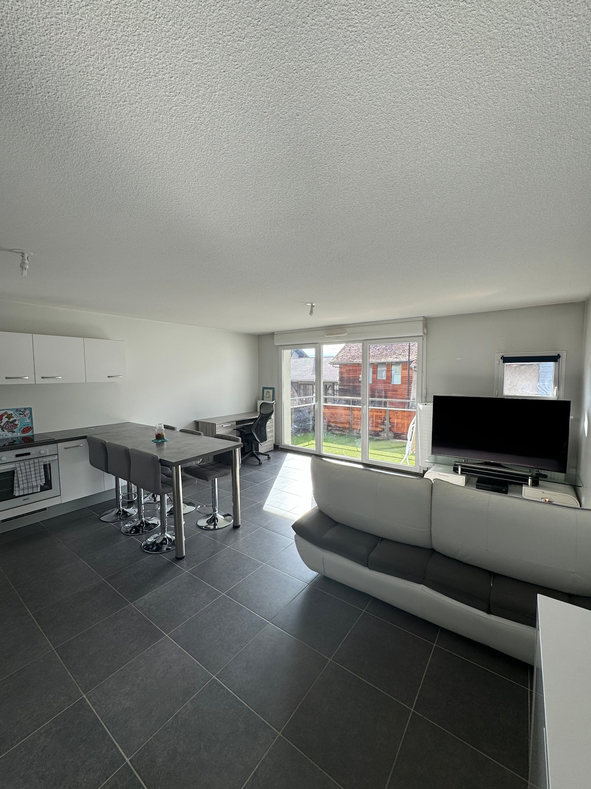 A modern living space is visible, featuring a sleek kitchen area with white cabinetry and a dining table with high stools. Natural light filters through large glass doors leading to an outdoor patio, while a television is positioned for viewing comfort.