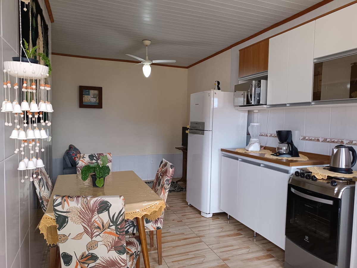 A well-equipped kitchen and dining area feature modern appliances, including a refrigerator and oven. A dining table is surrounded by floral-patterned chairs, providing a comfortable eating space. A ceiling fan enhances airflow in the room, which is brightly lit by natural light.