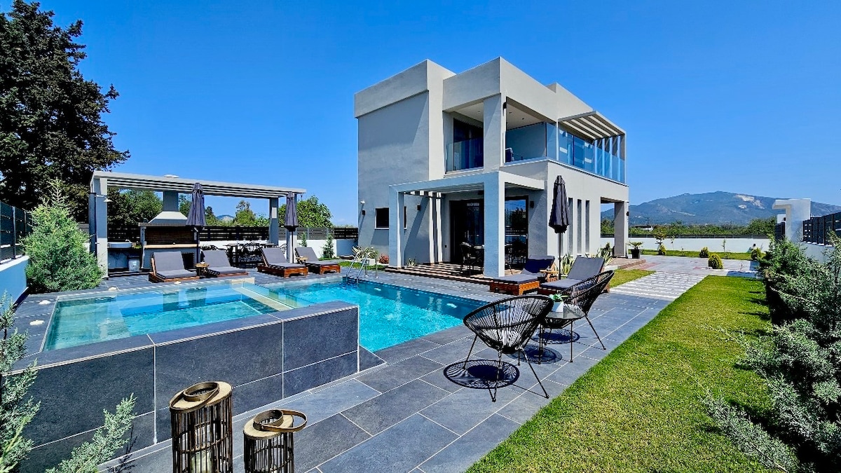 A newly constructed villa displays a modern design with large glass windows. A private pool and jacuzzi are positioned alongside a spacious patio, furnished with lounge chairs. Lush greenery surrounds the area, enhancing the serene outdoor environment with a clear blue sky above.