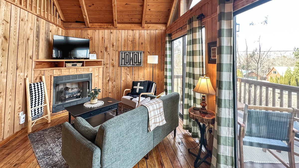 A rustic living area features wooden walls and a vaulted ceiling. A comfortable sofa is positioned across from a gas fireplace, with a flat-screen TV mounted above. Natural light floods the space through large windows, revealing a cozy reading nook with a lamp and chair.