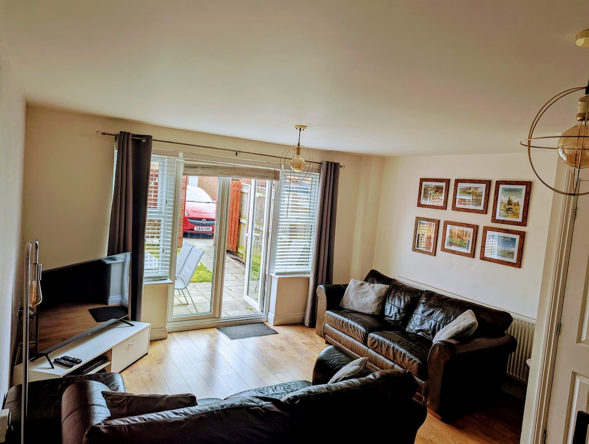 A welcoming living area is presented with two comfortable brown sofas and a flat-screen television. Large doors open to the garden, allowing natural light to fill the room. Framed images of various landscapes adorn the wall, enhancing the sense of space.