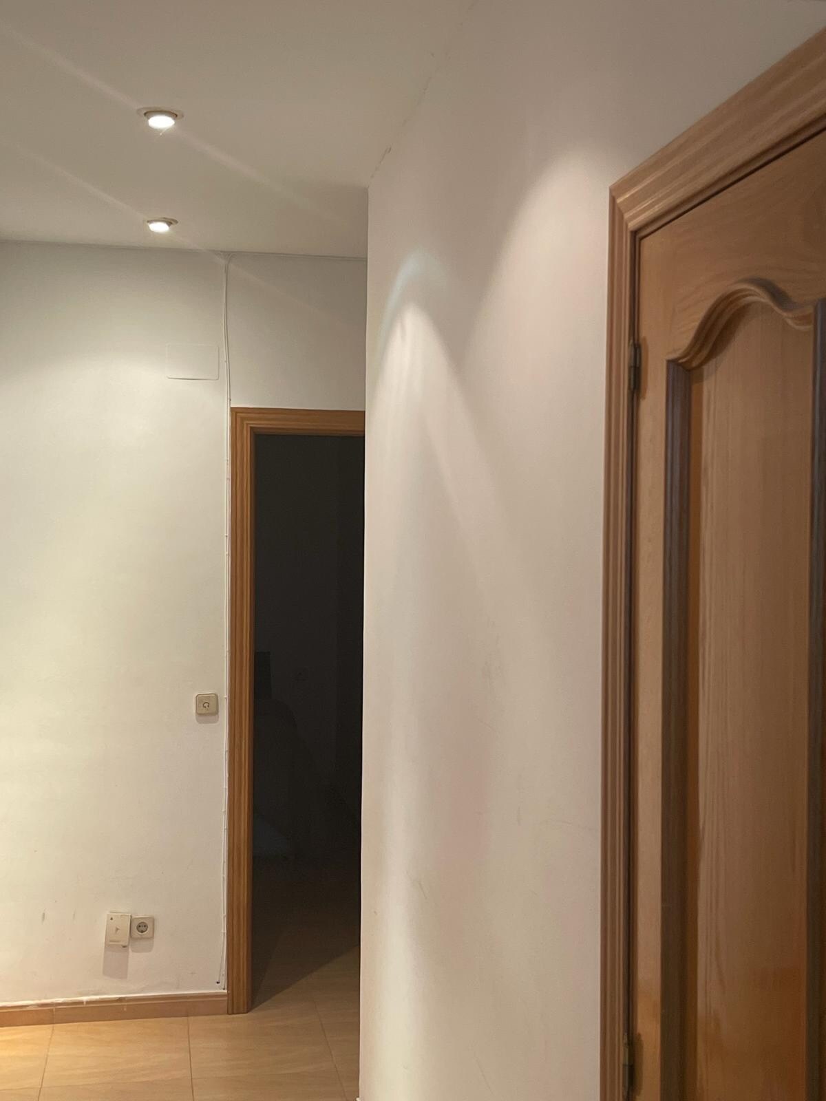 A corridor is visible, featuring a light-colored wall and a warm wooden door on the right. Ceiling lights provide illumination, highlighting the smooth surfaces of the walls and tiled flooring. An additional doorway is seen in the background, leading to another room.