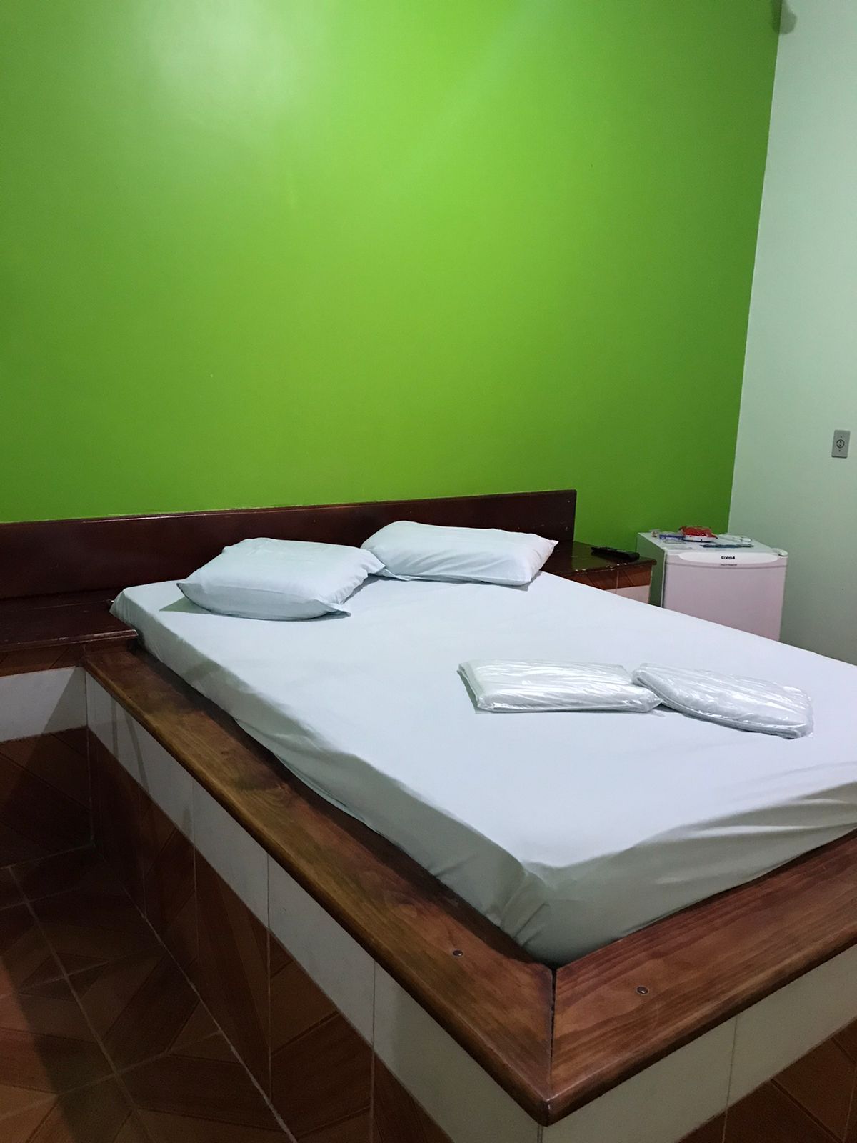 A simple sleeping area features a spacious bed with a light grey fitted sheet and two soft pillows. White towels are neatly placed on the bed. Bright green walls enhance the room’s fresh appearance, and a small fridge is visible nearby.
