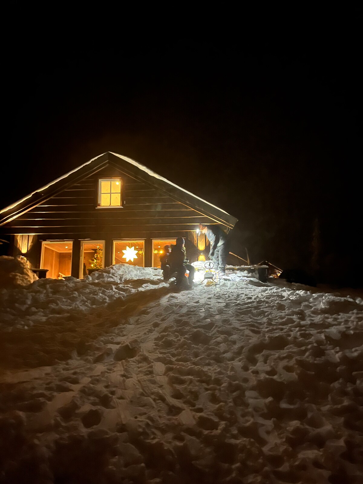 A cozy cabin is illuminated at night, featuring warm lights that highlight the wooden structure. Snow blankets the ground, and a path leads to the entrance. The front porch is adorned with seasonal decorations, creating a welcoming atmosphere amidst the winter landscape.