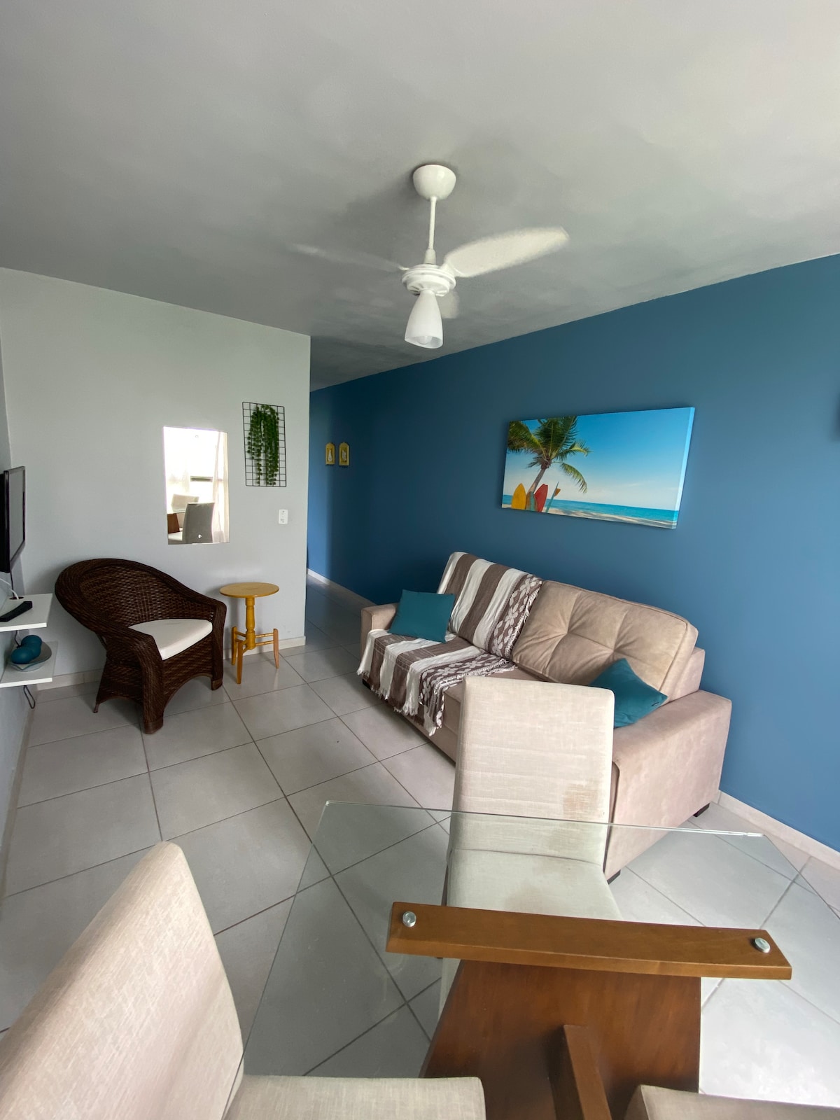 A bright living space features a beige sofa and a wicker chair. A small round table is positioned beside the sofa. The walls are painted in light blue, complemented by a palm tree artwork. A mirror reflects natural light, enhancing the room's openness.