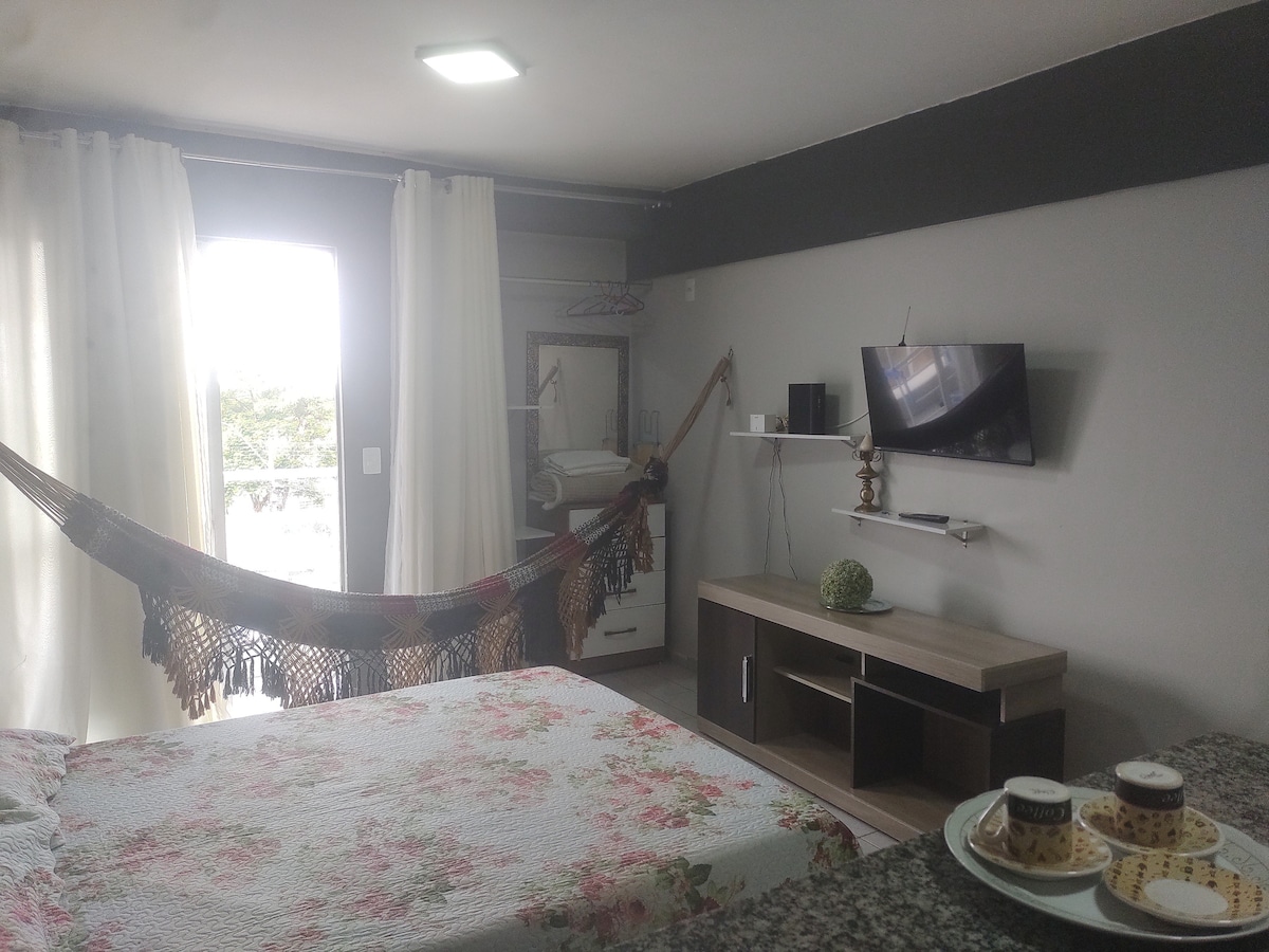 A sleeping area features a floral-patterned quilt covering a spacious bed. A hammock hangs between two points, and a television is mounted on the wall. Natural light enters through glass doors, adorned with sheer white curtains, leading to a small outdoor space.