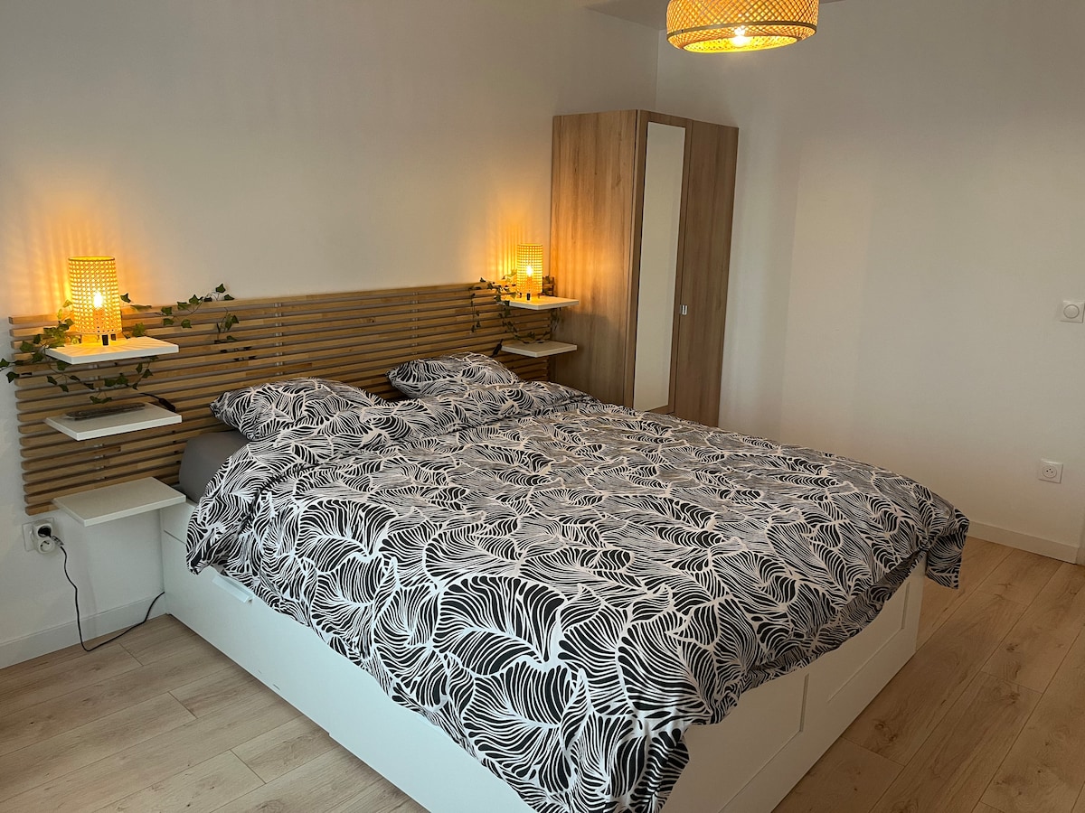 A spacious bedroom features a large bed adorned with a patterned duvet cover. Wooden shelving and a wardrobe provide storage options. Soft lighting from two lamps adds ambiance, while greenery subtly decorates the headboard.