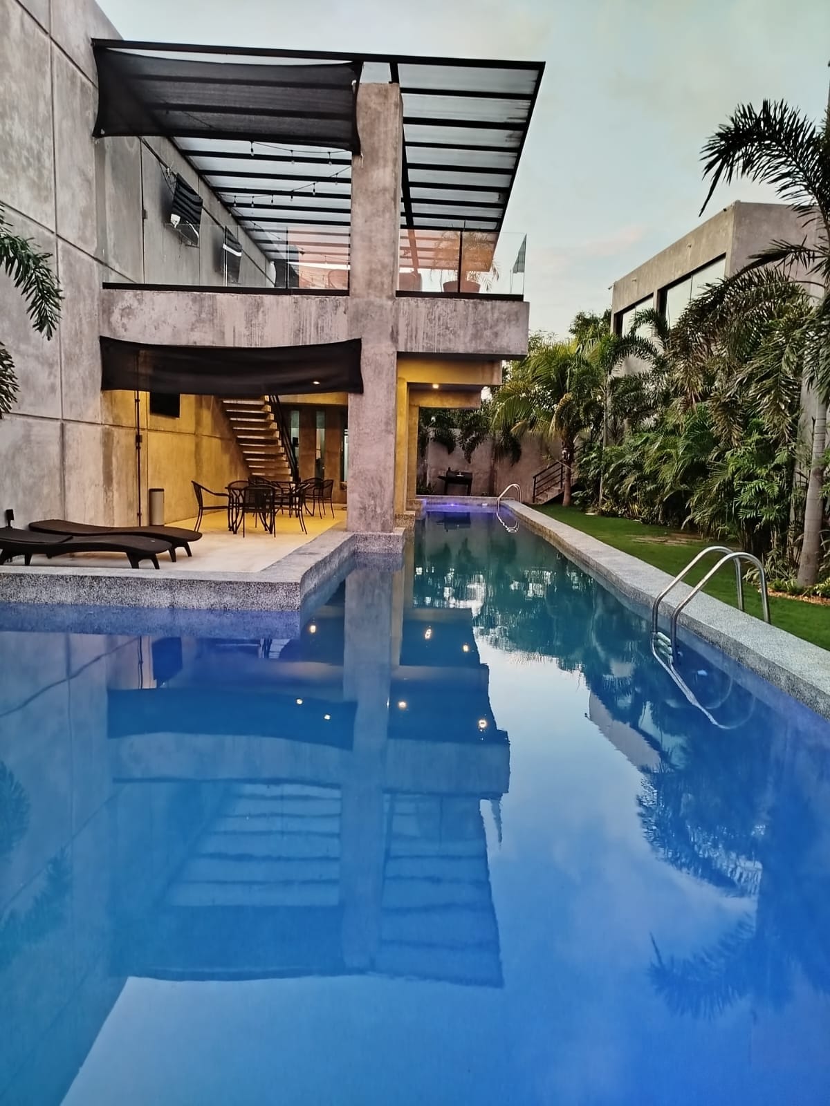 A sparkling pool reflects the evening sky, surrounded by lush greenery. The area features a modern building with open spaces and a staircase leading to an upper level. Comfortable seating arrangements are present nearby, creating an inviting outdoor atmosphere.
