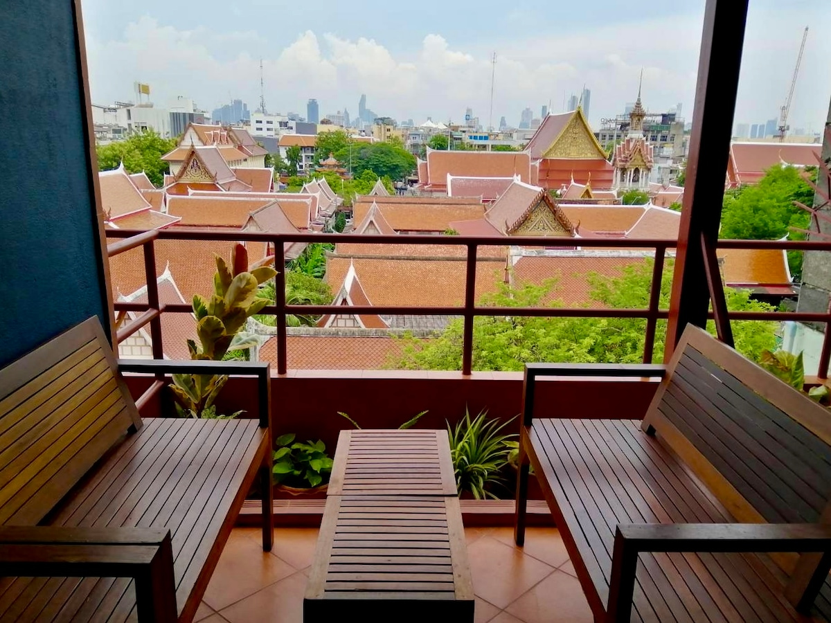 A private balcony showcases a view of traditional Thai rooftops and the distant skyline. Two wooden chairs and a small table are positioned on the terrace, framed by greenery and city structures. Soft natural light enhances the outdoor space.