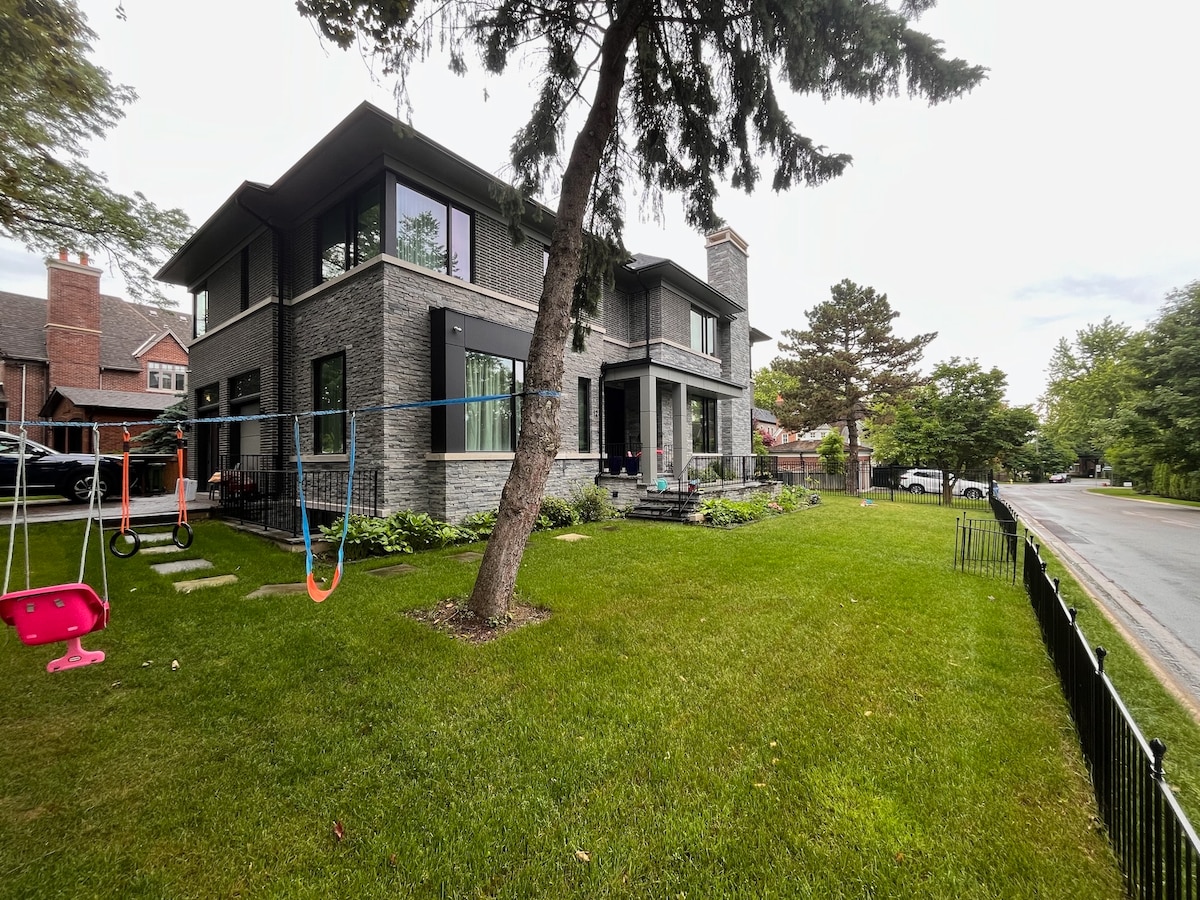 A modern house showcases large windows and a stone exterior. The front yard is well maintained with green grass and flowerbeds. A swing set and slide are visible in the yard, adding a playful touch to the inviting outdoor space.
