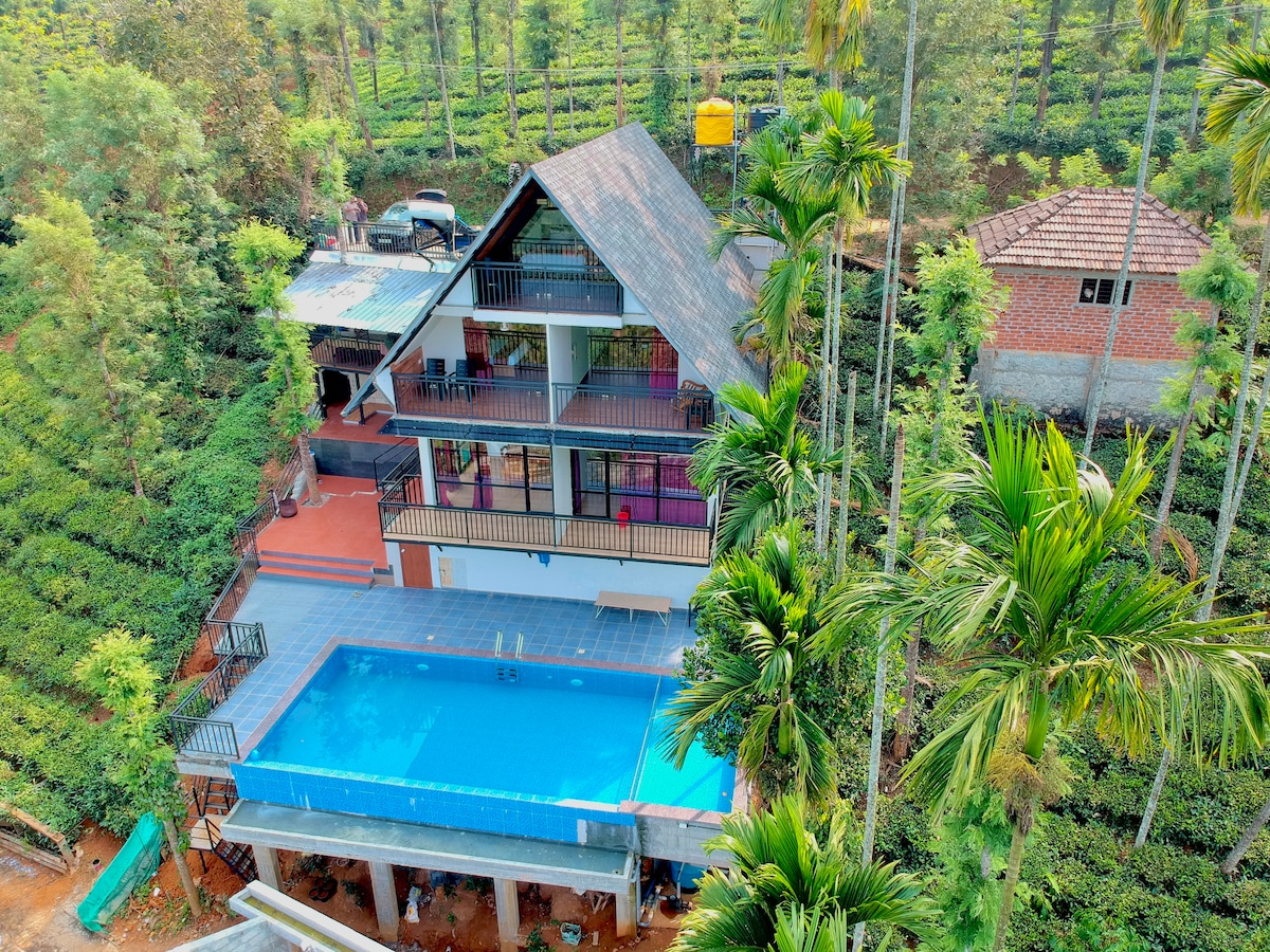 A multi-level villa is nestled among lush greenery, providing a view of a private pool on the lower level. The expansive outdoor area features a spacious deck, surrounded by palm trees and distant hills. The structure exhibits a modern design with multiple balconies.