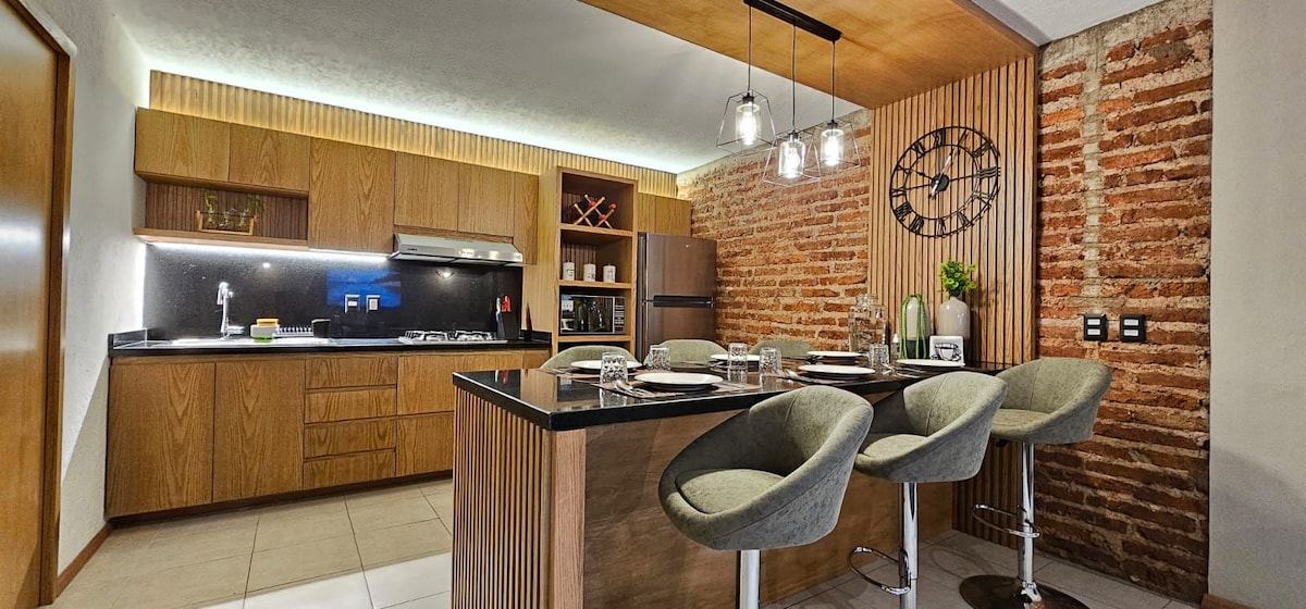 A modern kitchen features wooden cabinets and a black countertop with four cushioned bar stools. A stove, oven, and sink are visible, complemented by brick accent walls and soft overhead lighting, enhancing the space's contemporary design.