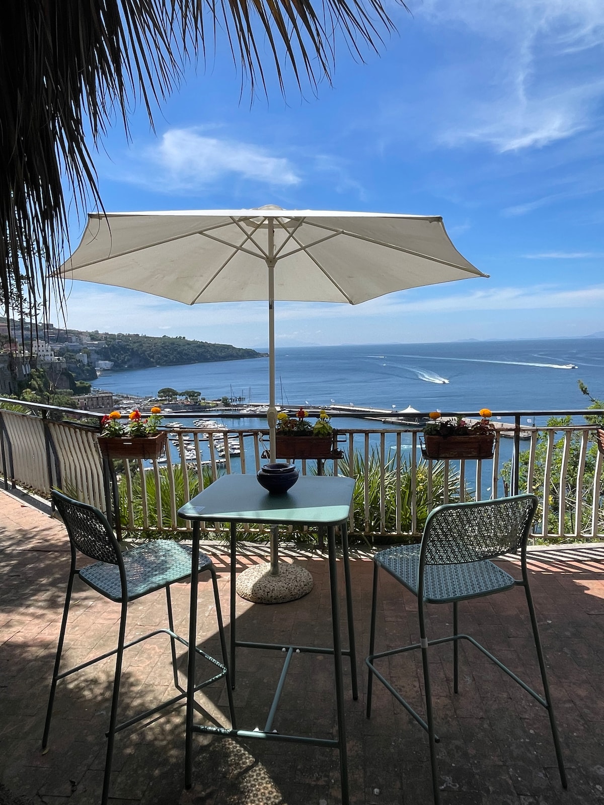 A shaded outdoor seating area is seen, featuring a small round table and two high chairs. An umbrella provides shade over the table, while vibrant flowers can be seen along the railing, framing a panoramic view of the sea and coastline under a clear blue sky.