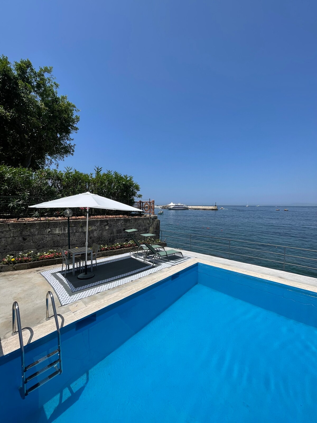 A tranquil infinity pool is seen overlooking the sea, with inviting seating under a large umbrella. Lush greenery surrounds the area, enhancing the feeling of serenity. The clear blue water of the pool reflects the unobstructed sky, creating a seamless transition to the ocean beyond.