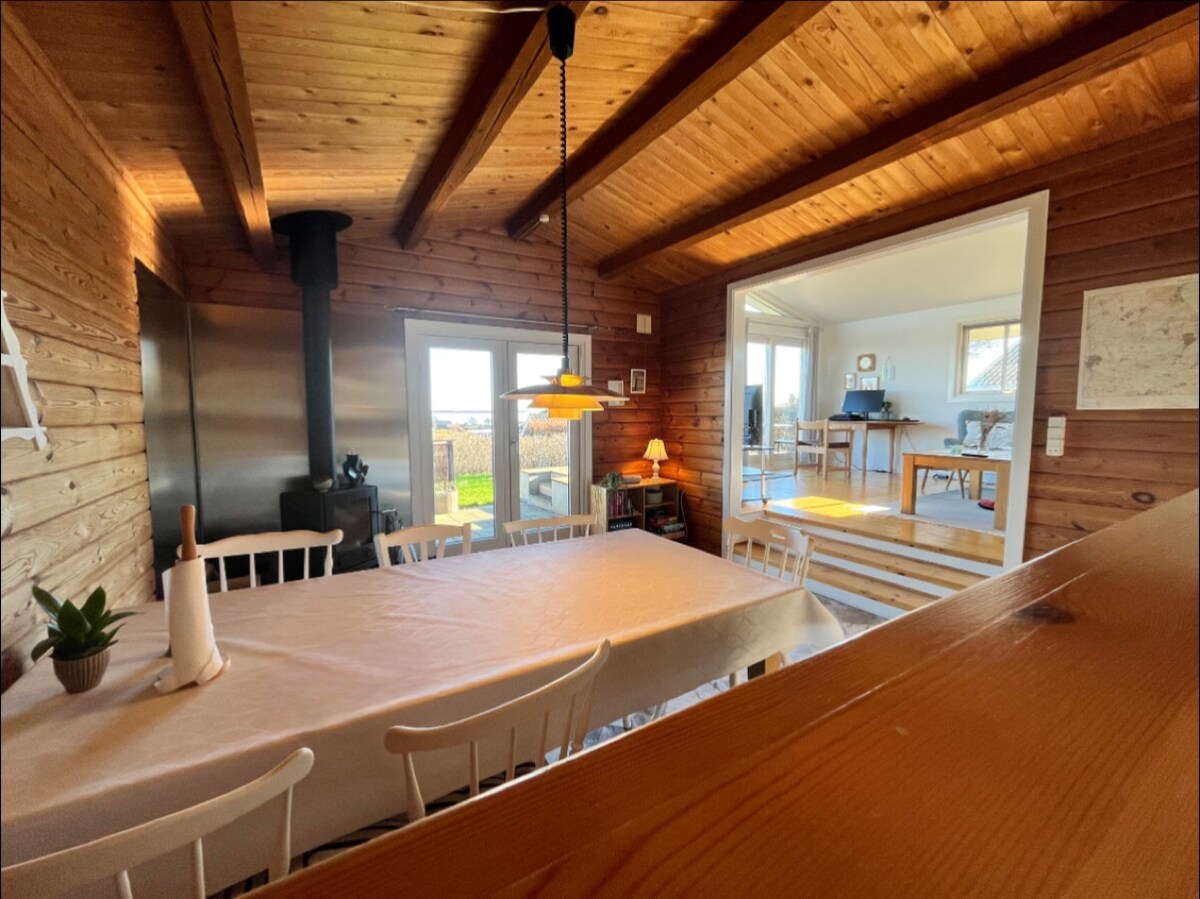 A spacious dining area is illuminated by natural light, featuring a large table surrounded by several white chairs. A wooden ceiling with exposed beams complements a wood-burning stove, while a doorway leads to an adjoining room with additional seating and outdoor views.