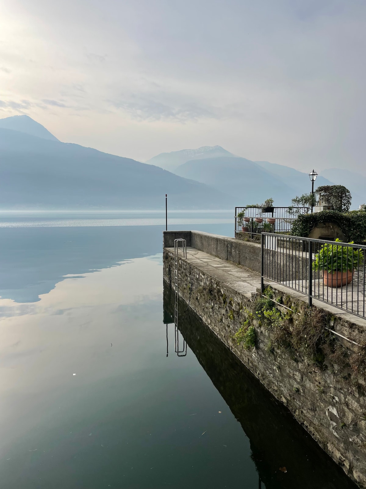 A serene view of a lake is captured, with mountains reflected across the still water. A stone wall borders the waterfront, featuring a railing and a planter with greenery. Soft blue hues dominate the scene, creating a calm atmosphere.