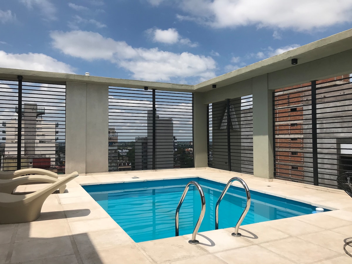 A rooftop terrace is featured with a rectangular swimming pool surrounded by a tiled deck. Lounge chairs are arranged nearby, and large slatted panels provide both privacy and views of the cityscape beyond. The sky is partly cloudy, adding to the spacious feel.