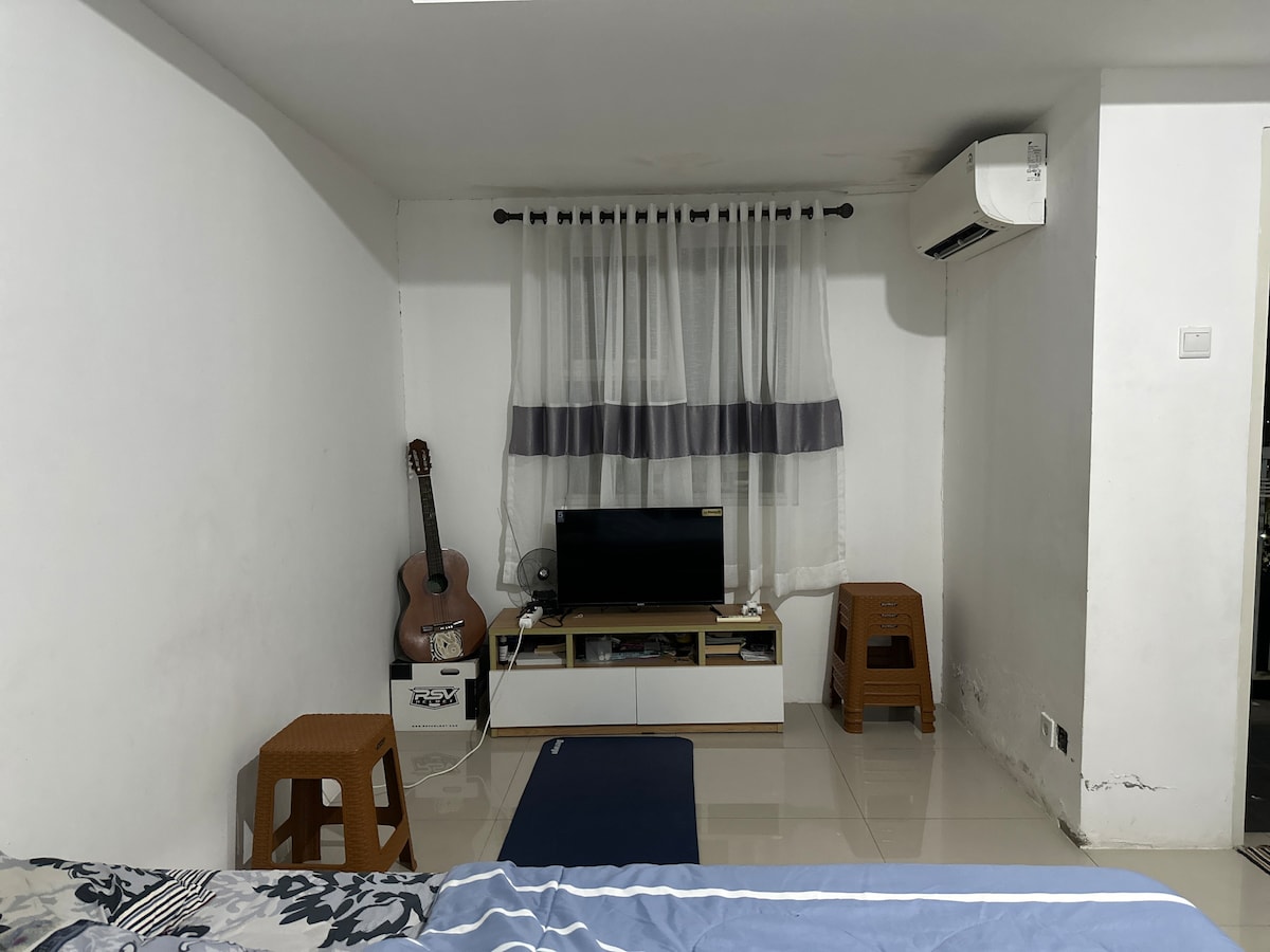 A compact living area is presented, featuring a low television stand with a screen, a fan, and a placed guitar. Two small wooden stools are positioned nearby, while a blue mat lays on the glossy floor. Natural light filters through sheer curtains adorning the window.