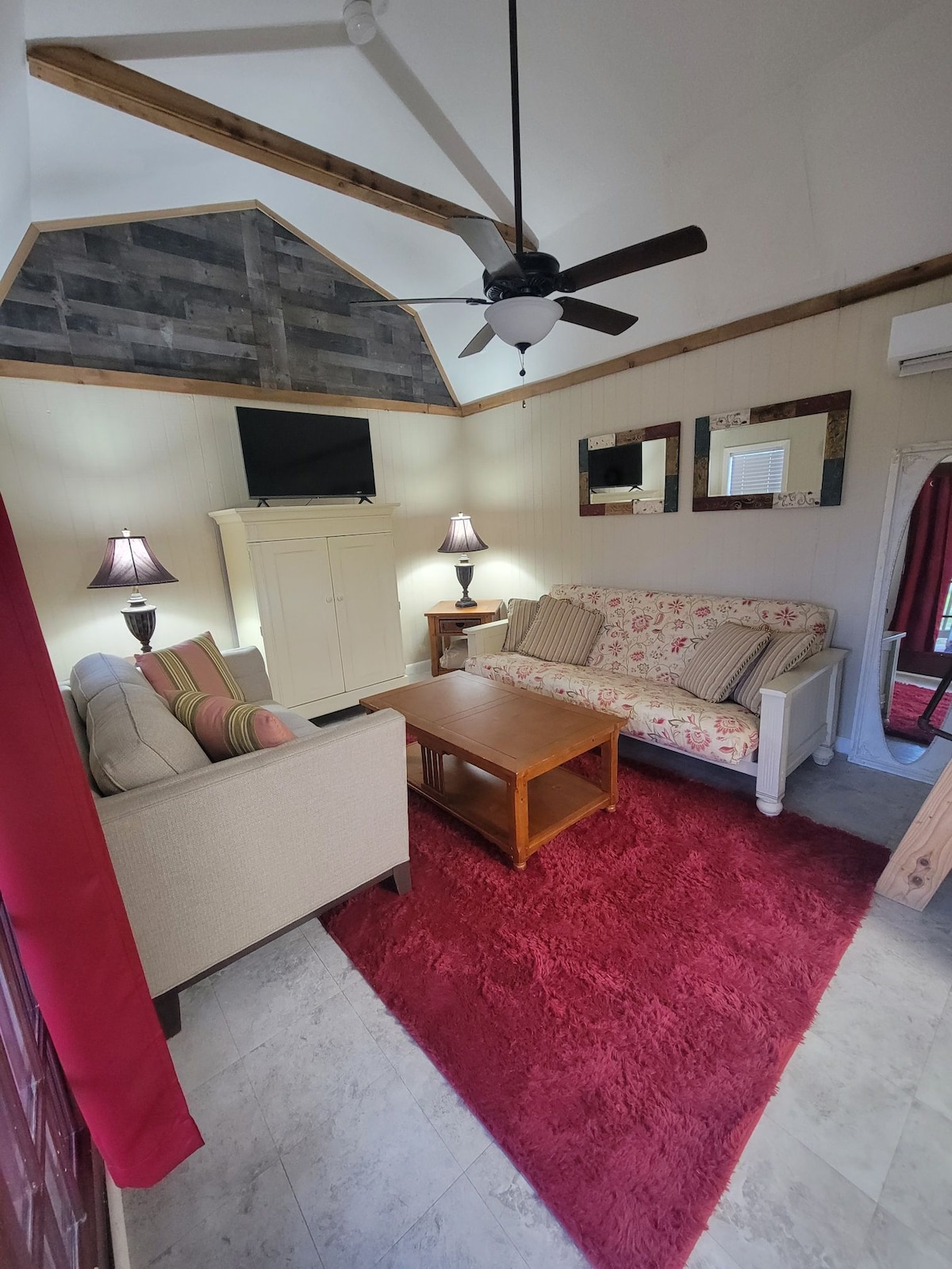 A cozy living area is presented with two sofas, one adorned with floral patterns. A wooden coffee table sits at the center, complemented by softly glowing table lamps on either side. A television is mounted on the wall, enhancing the room's functionality.