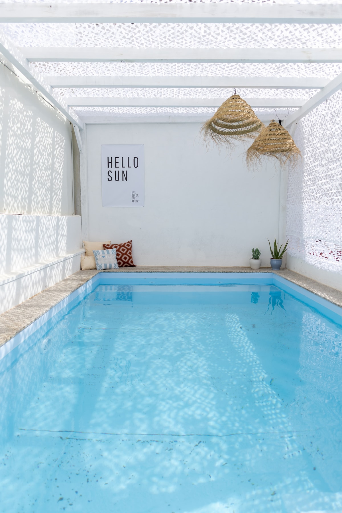 A private saltwater pool is featured within a sheltered area, illuminated by natural light. The pool is surrounded by a simple stone border. Decorative elements include two hanging light fixtures and a small plant on the ledge, with a wall display that reads 'HELLO SUN.'