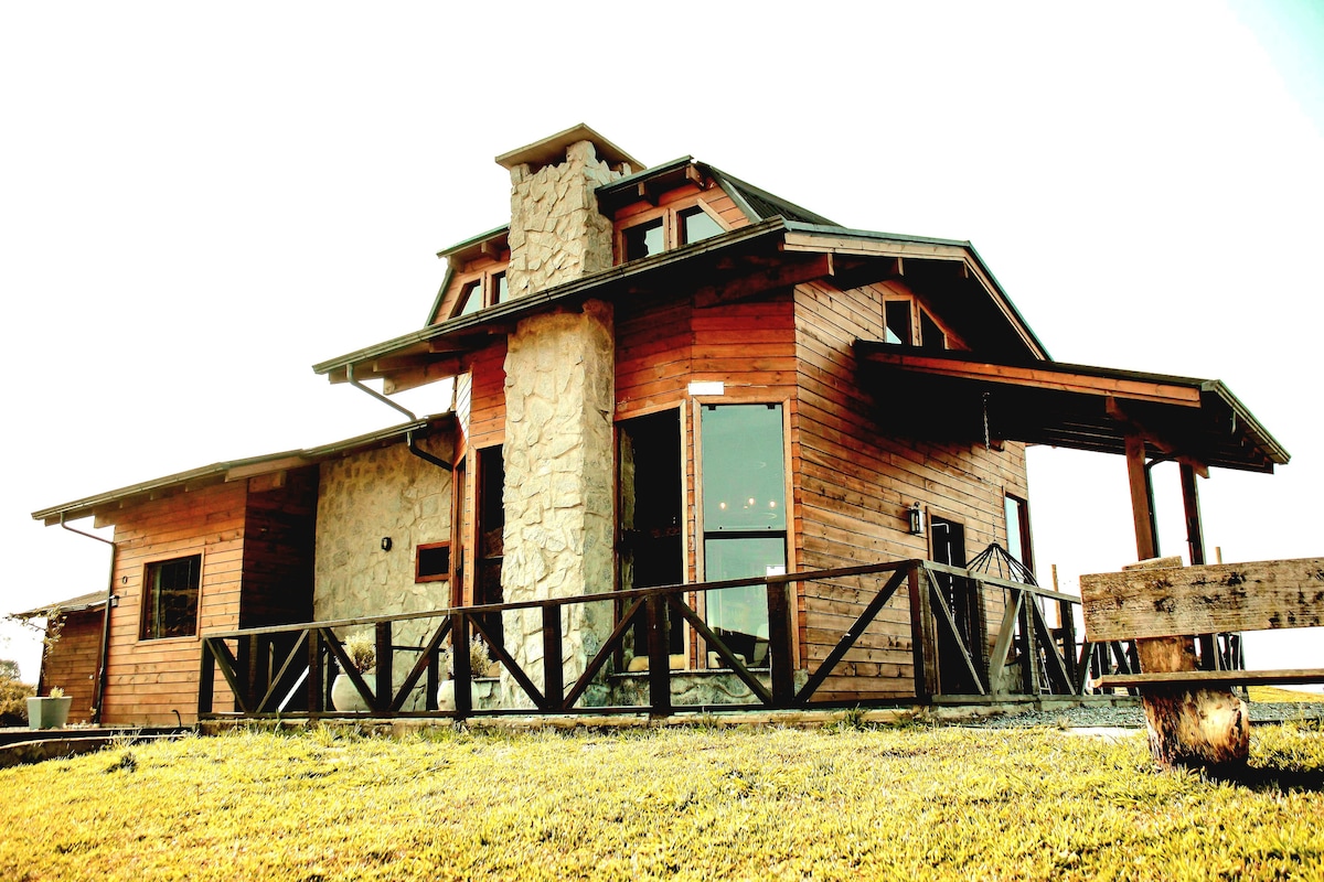 A charming wooden house is presented, featuring a stone accent wall and a large front balcony. Expansive glass doors invite natural light, while the sloped roof contributes to the rustic appeal. Surrounded by a grassy area, the home showcases a scenic countryside setting.