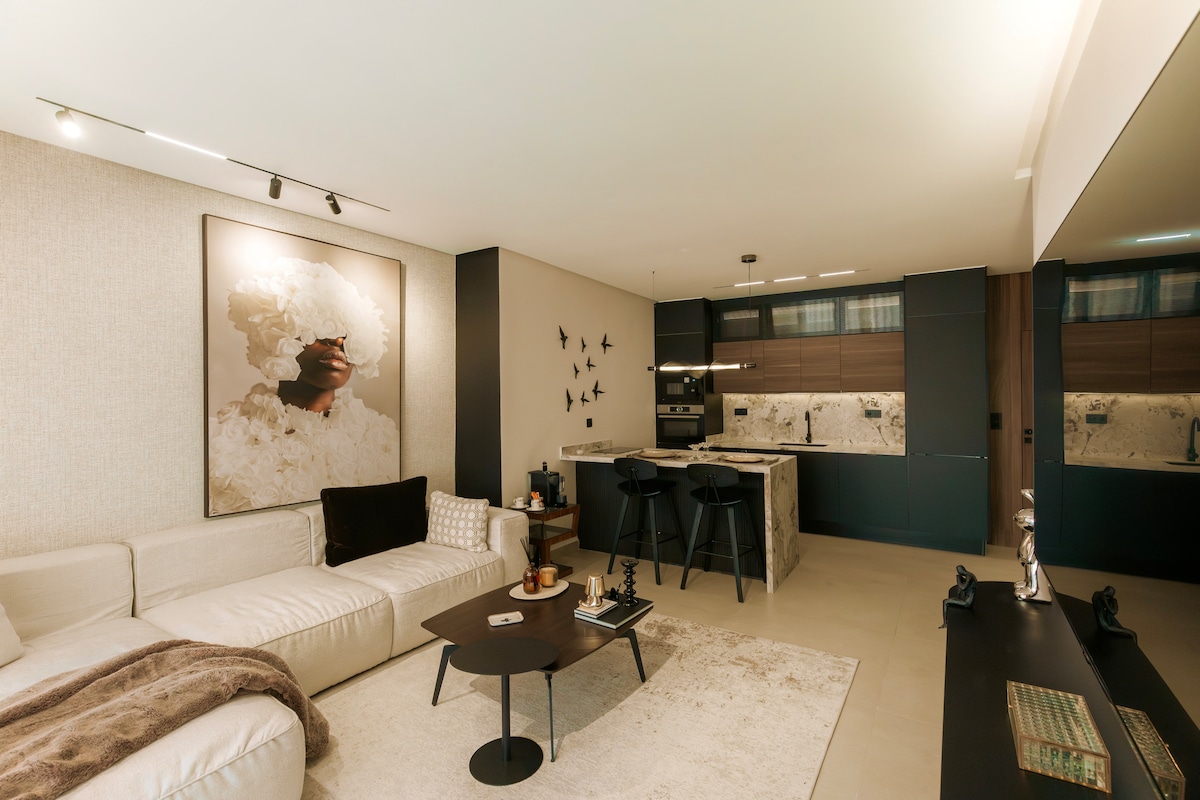 A modern living area features a comfortable sofa draped with a cozy blanket, accompanied by a sleek coffee table. The open kitchen, visible in the background, boasts elegant cabinetry and contemporary appliances. A striking piece of wall art enhances the overall sophisticated decor of the space.