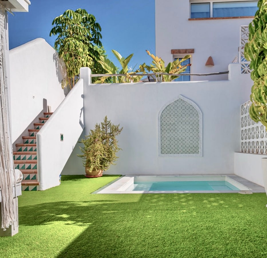 A private pool is nestled within a grassy area, surrounded by lush greenery. A staircase featuring patterned tiles leads to an upper level on the left. The white walls and decorative arch in the rear create a serene atmosphere.