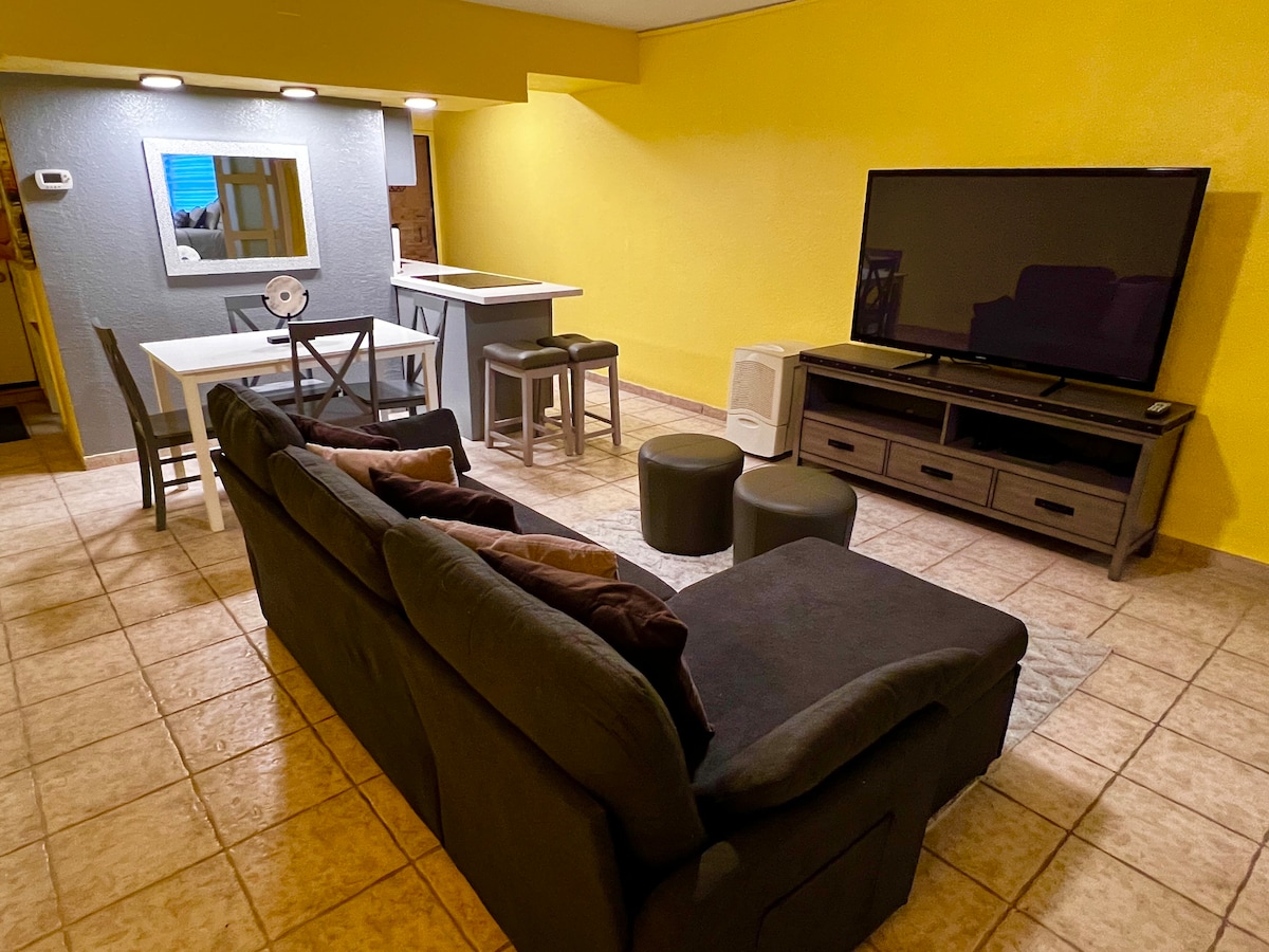 A cozy living area is featured, characterized by a dark sofa with throw pillows. A large television is mounted on a wooden console. A dining space with a table and gray chairs is seen in the background, complemented by warm yellow walls.