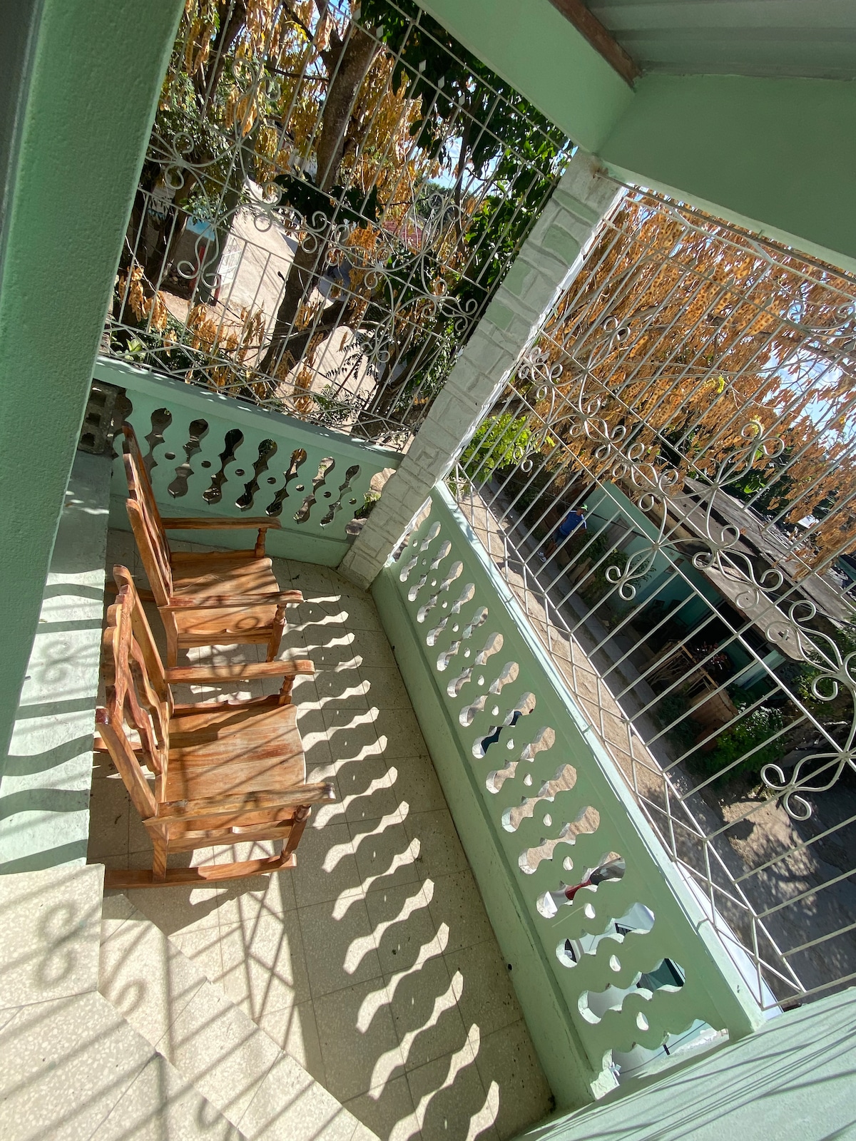 A cozy balcony features two wooden chairs positioned to enjoy the view. Intricate iron railings allow natural light to cast playful shadows on the tiled floor, surrounded by greenery from nearby trees.