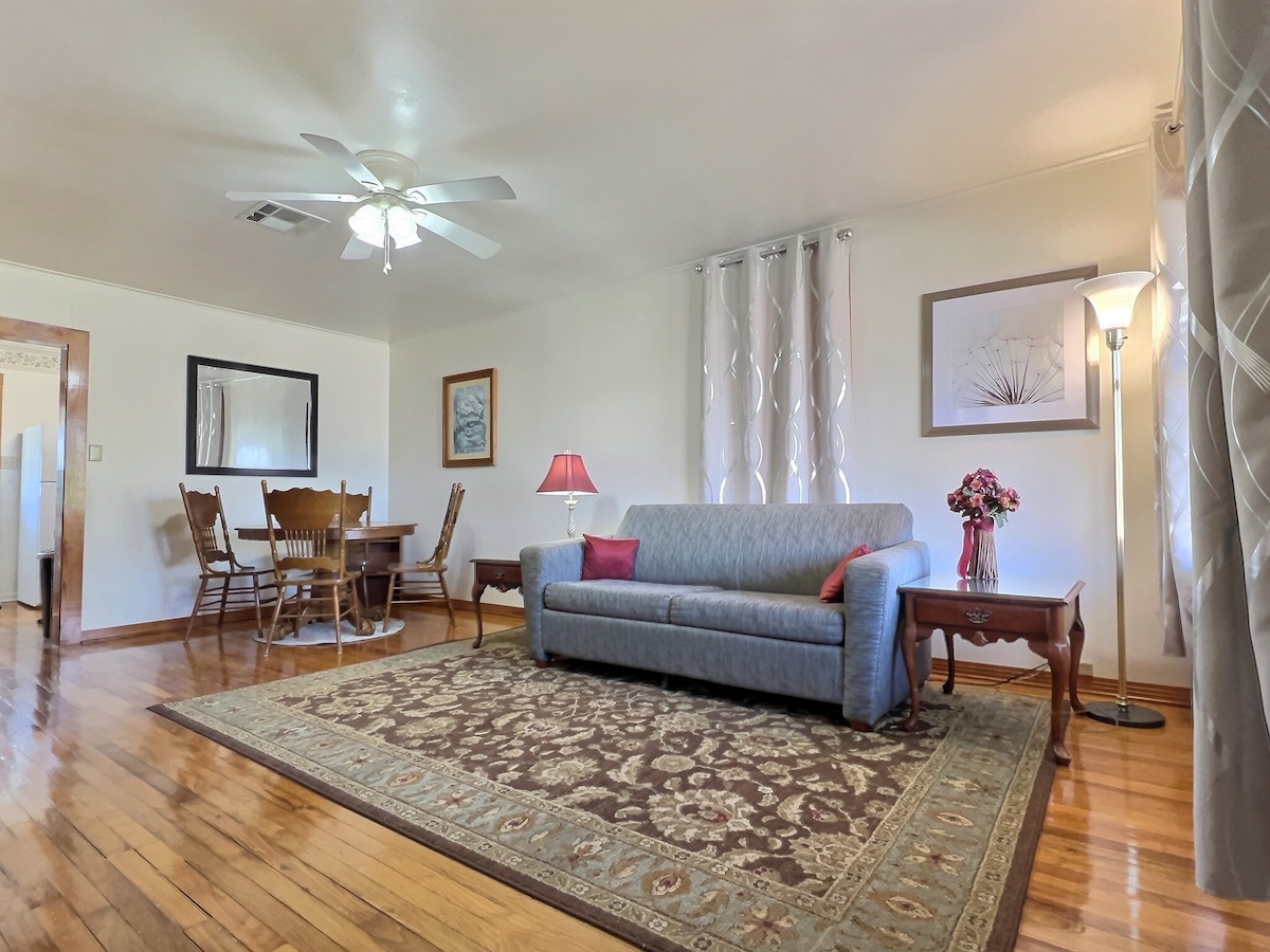 The living area features hardwood floors and a comfortable pullout sofa, complemented by a decorative area rug. A dining table with chairs is positioned nearby, while natural light filters through the window drapes. A lamp and floral arrangement add subtle touches to the space.