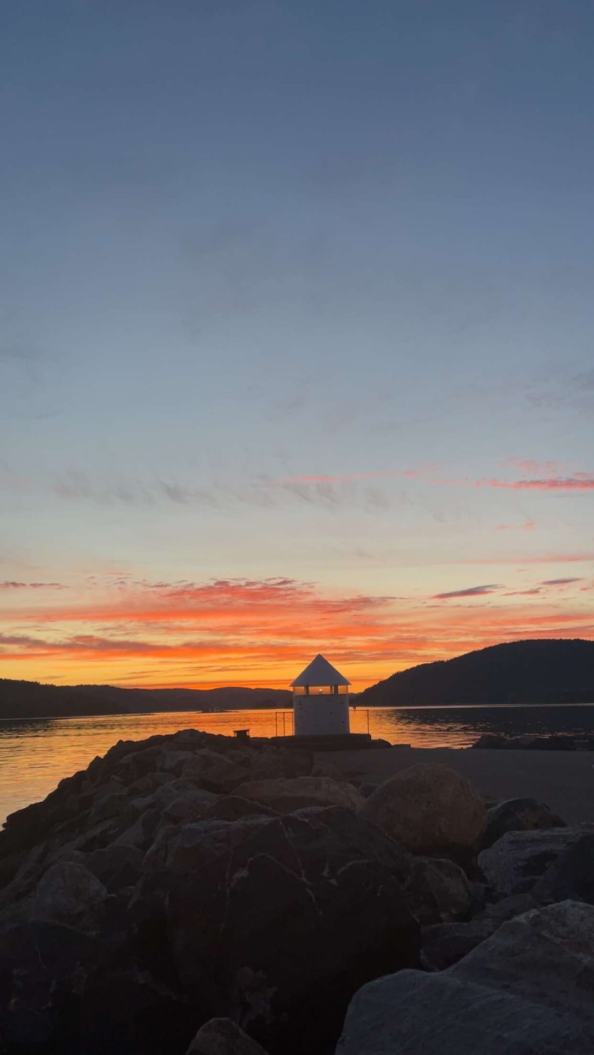 A sunset view features a small structure atop rocks, silhouetted against a vibrant sky filled with hues of orange, pink, and blue. The water reflects the colorful sky, and silhouettes of distant hills can be seen in the background.