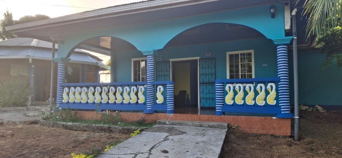A colorful exterior of a building is highlighted by blue walls and contrasting white and yellow decorative elements on the porch railing. Arched entryways create an inviting atmosphere, while the surrounding vegetation adds a touch of nature to the setting.