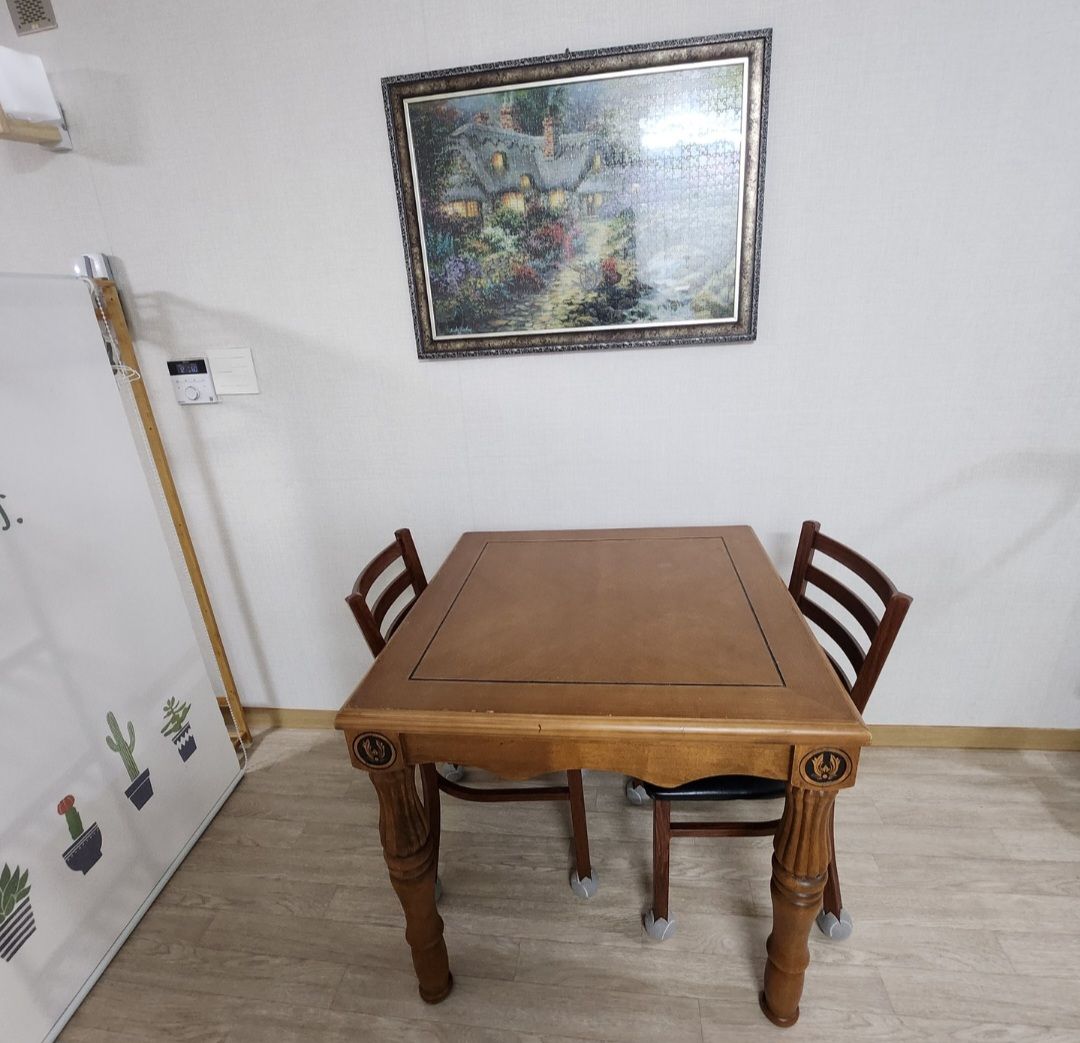 A wooden table is positioned centrally in the image, accompanied by two wooden chairs. A framed artwork adorns the wall above, depicting a serene landscape. The floor features light-colored tiles, contributing to a clean and simple ambiance.
