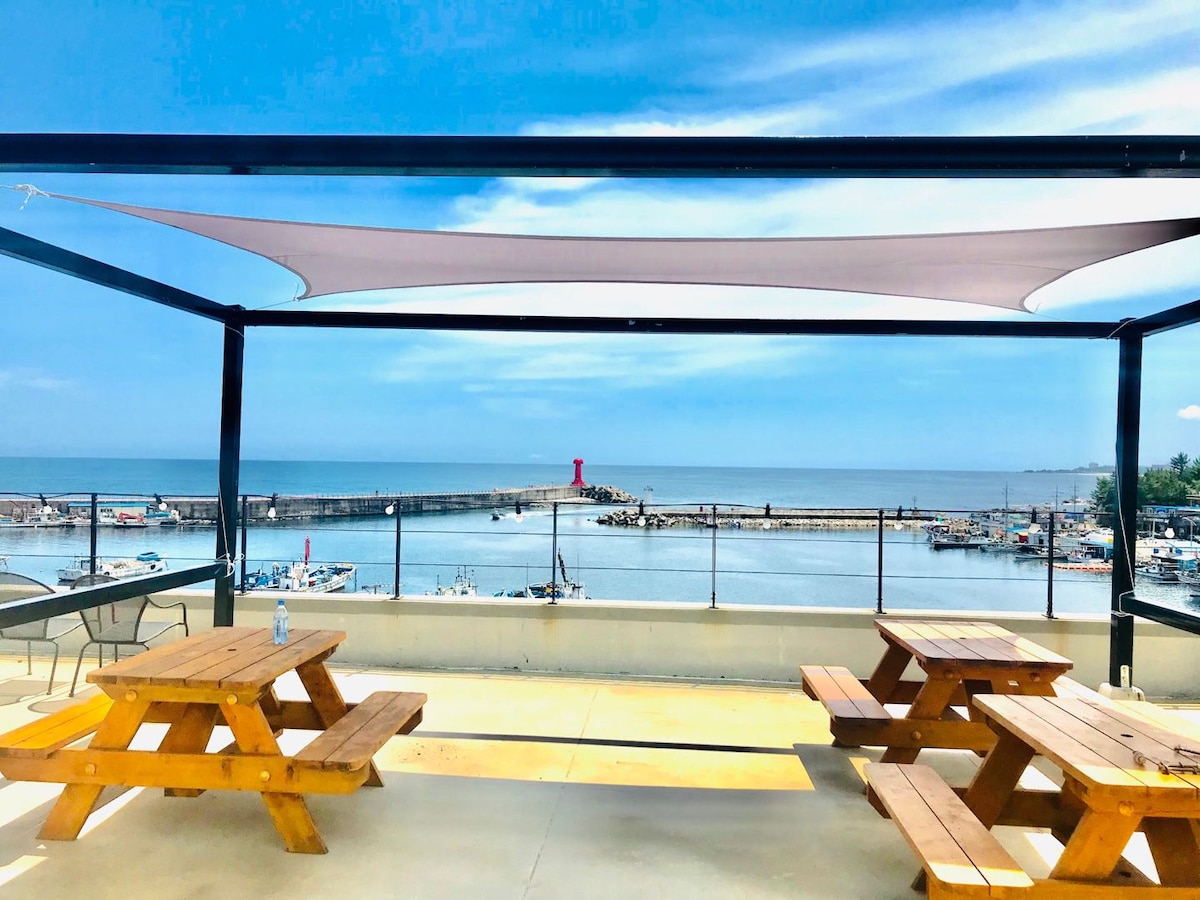 A spacious rooftop terrace features wooden picnic tables shaded by a fabric canopy. The view includes a vibrant harbor with a red lighthouse, surrounded by boats. Light clouds drift across a blue sky, enhancing the serene coastal atmosphere.