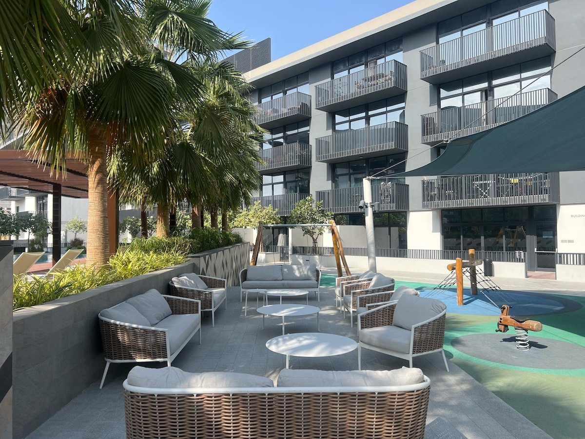 An outdoor seating area is presented with multiple lounge chairs and a few tables, surrounded by lush greenery and palm trees. The modern building facade is visible in the background, creating a relaxed atmosphere. A shaded area offers protection from the sun.