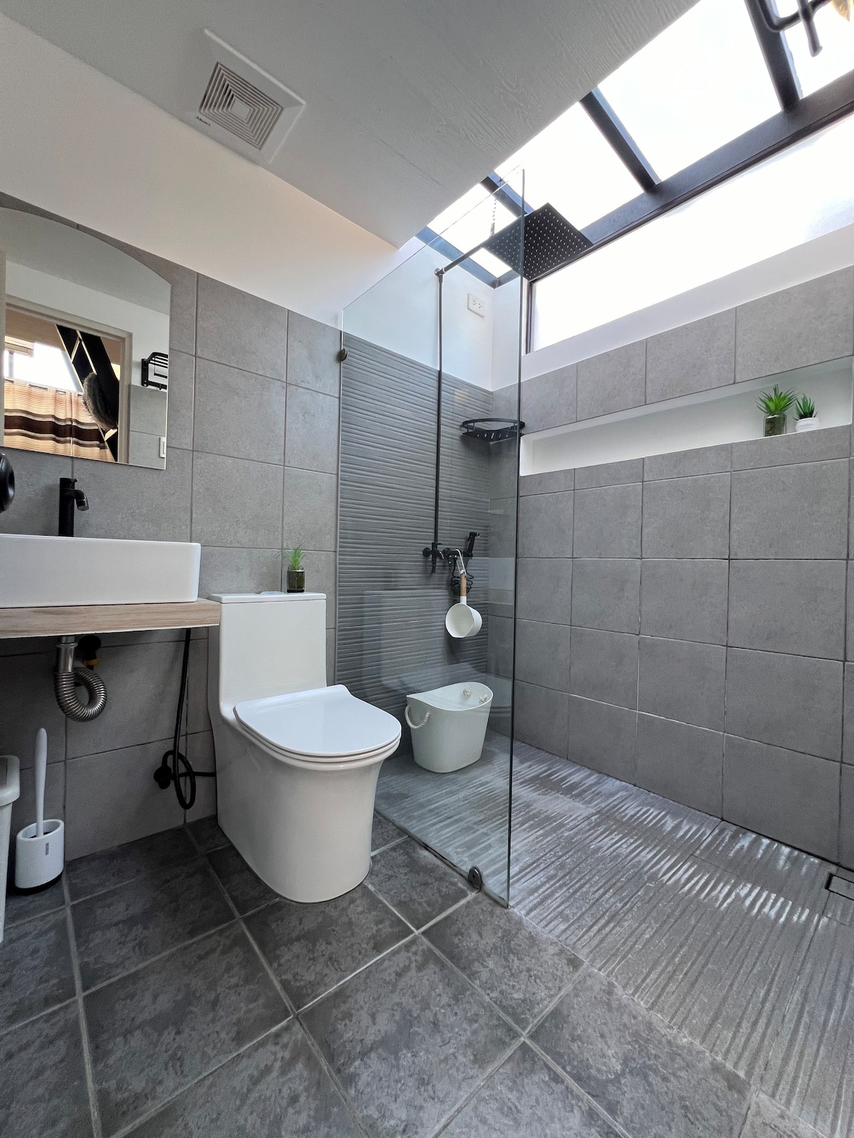 A modern bathroom is showcased, featuring gray tiled walls and floor. A glass shower enclosure is equipped with a rainfall shower head. A floating vanity holds a stylish sink and a mirror, alongside small plants for subtle decor. Natural light is let in through a skylight overhead.