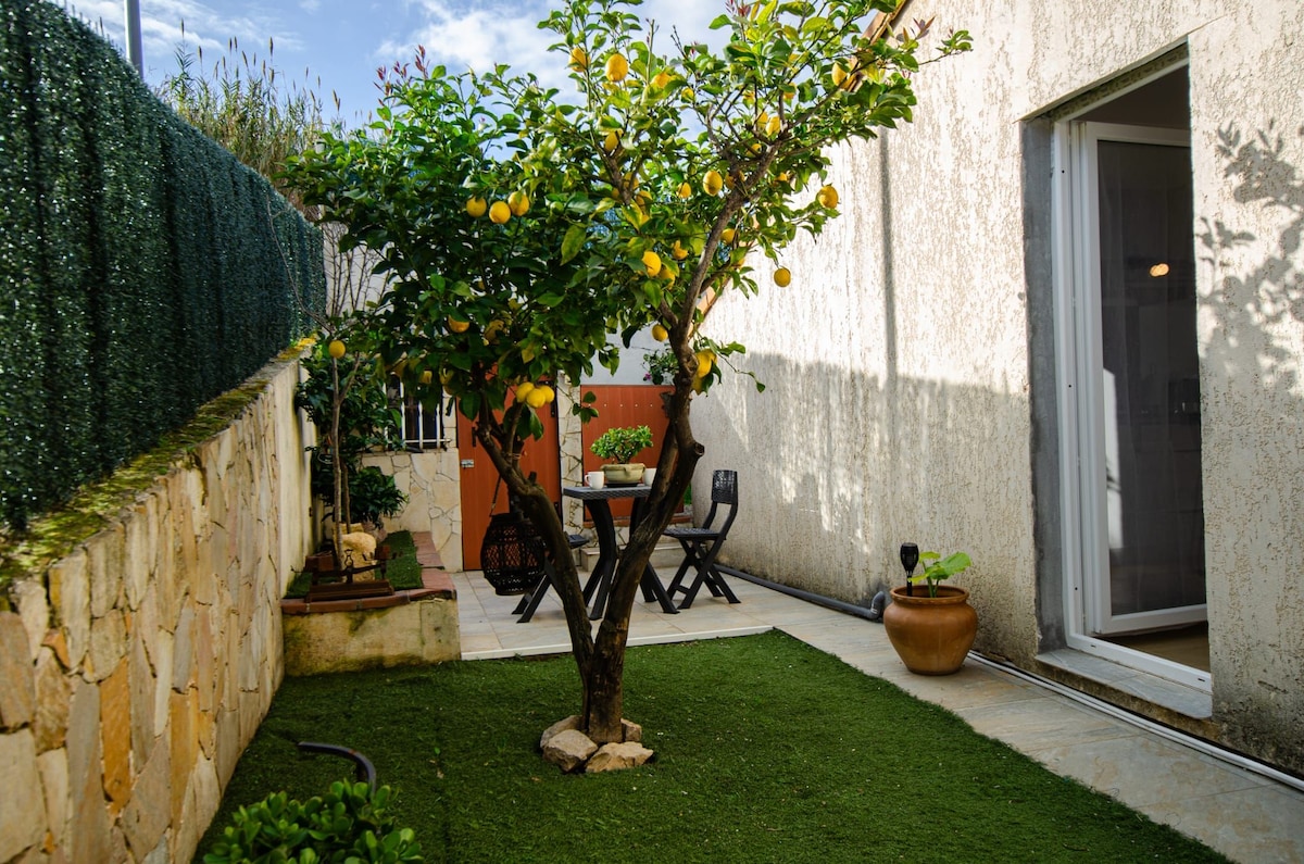 A lemon tree stands in the center of a green garden, surrounded by neatly trimmed grass. A small outdoor dining table with chairs is positioned nearby, offering a cozy space for meals. Natural light highlights the serene atmosphere of the area.