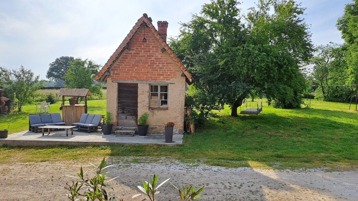 A charming rustic building features a brick facade and wooden door, situated amidst green grass and trees. A spacious terrace with lounge chairs is present in front, along with planters. A swing can be seen in the background, creating a serene outdoor setting.