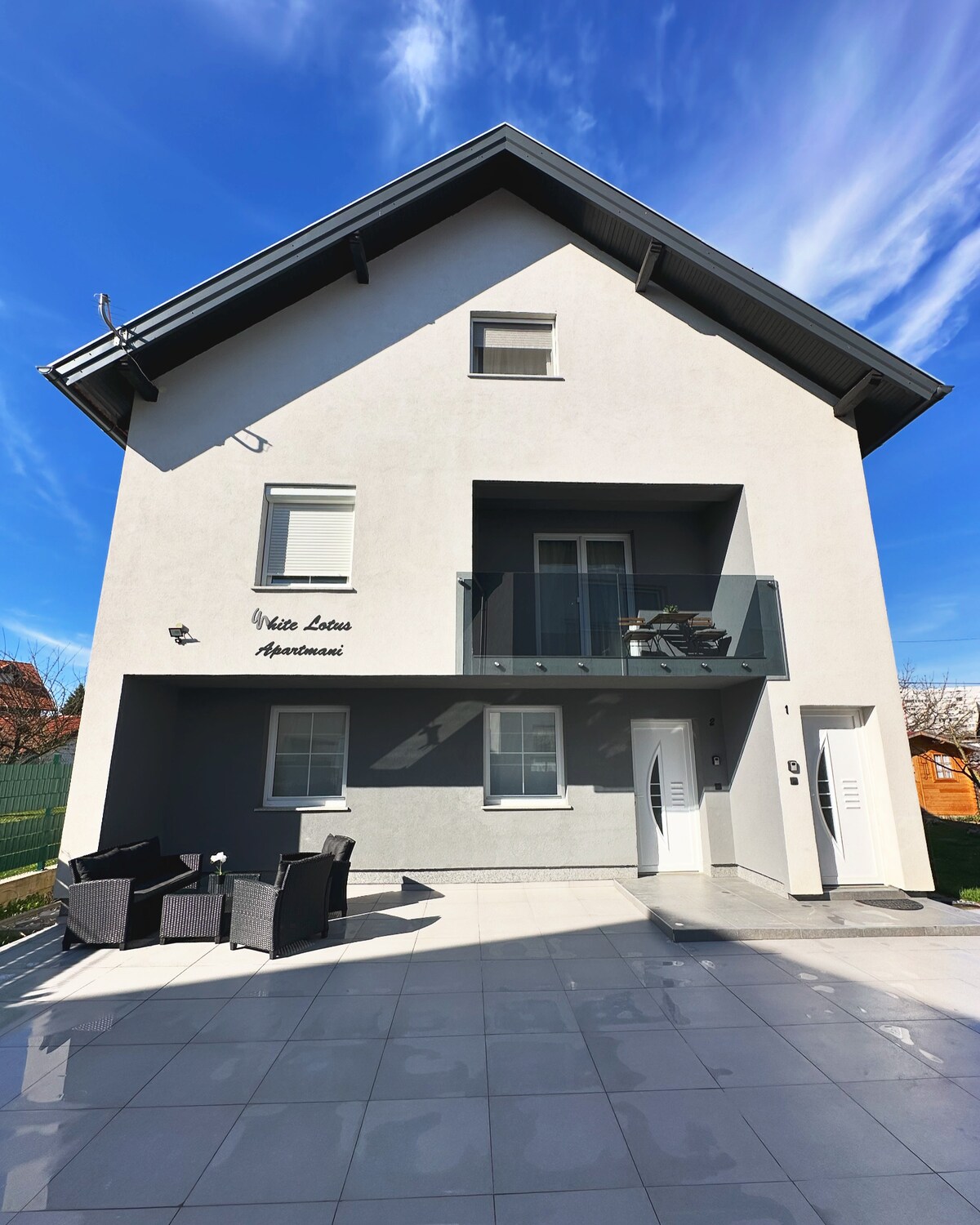 The exterior of the White Lotus apartment building is depicted, showcasing a modern design with a two-story structure. A small balcony is visible on the upper floor, while a seating area with wicker chairs is arranged on the ground level. Clear blue skies enhance the image.