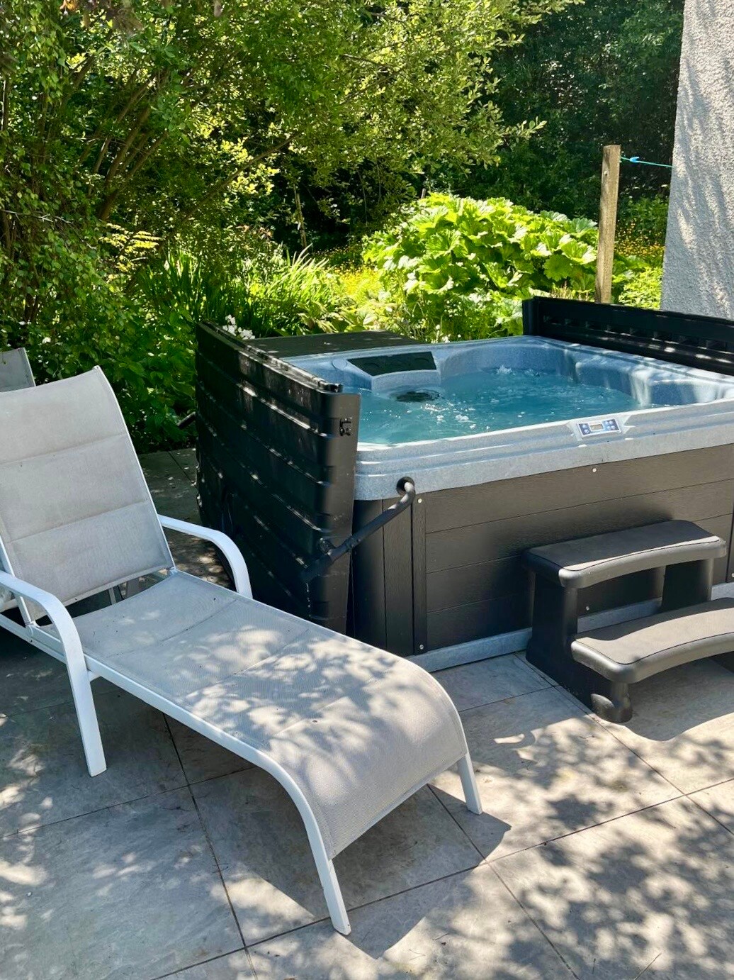 A hot tub is positioned on a spacious patio surrounded by lush greenery. A white lounge chair and a small step stool sit nearby on the gray stone tiles, offering a place to relax. The area is well-maintained, creating a serene outdoor setting.