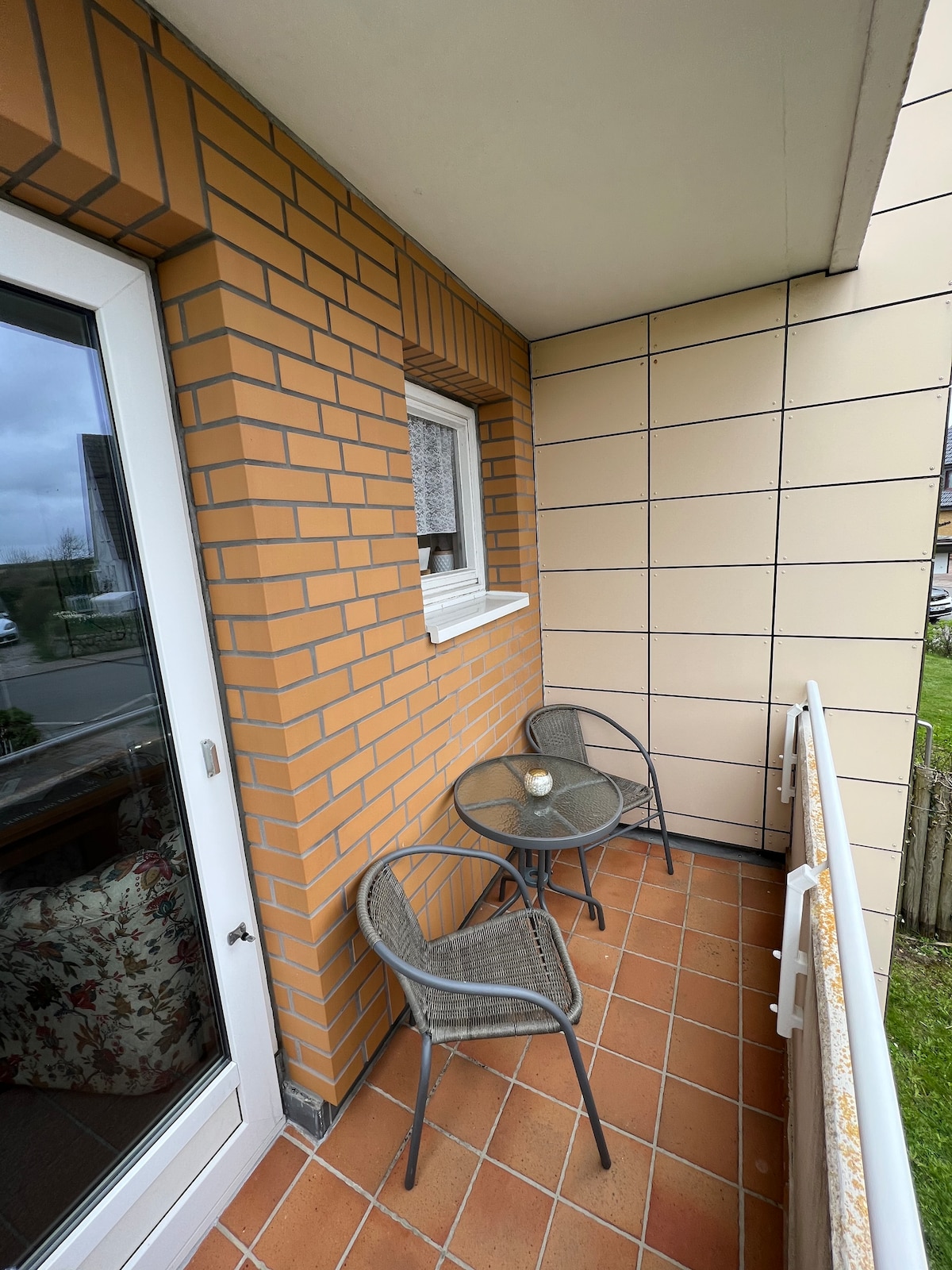 A small balcony is presented with warm, patterned brick walls and a tiled floor. Two wicker chairs are positioned around a round table. A glass candle holder sits atop the table, providing a simple decorative touch. The view extends over a green space beyond the railing.