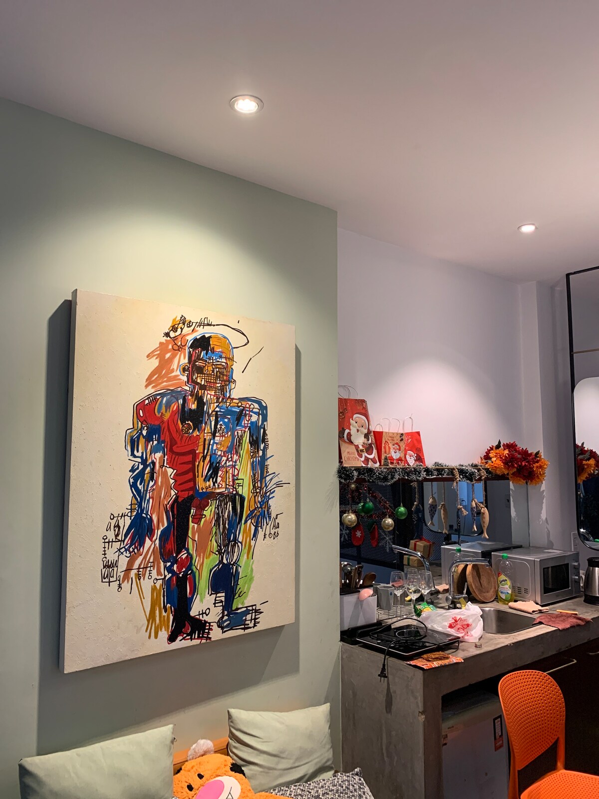 A vibrant abstract painting dominates the wall, featuring bold colors and expressive lines. A compact kitchenette is visible with a microwave and kitchen items displayed. Decorative accents, including festive ornaments, are arranged on shelves above the workspace, contributing to the lively atmosphere.