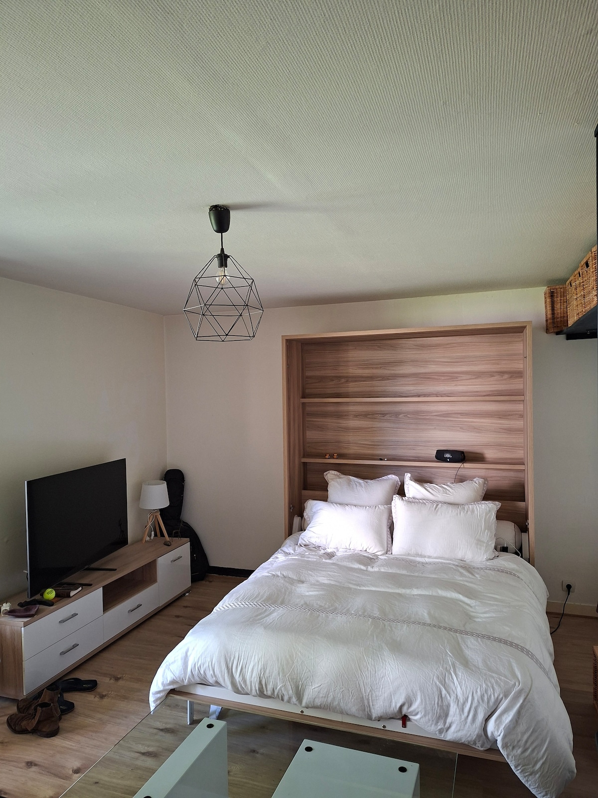 A well-arranged studio features a compact sleeping area with a spacious bed dressed in white linens. A wooden wall unit serves as a backdrop, complemented by a stylish geometric light fixture above. A television is positioned on a low, minimalist console across from the bed.