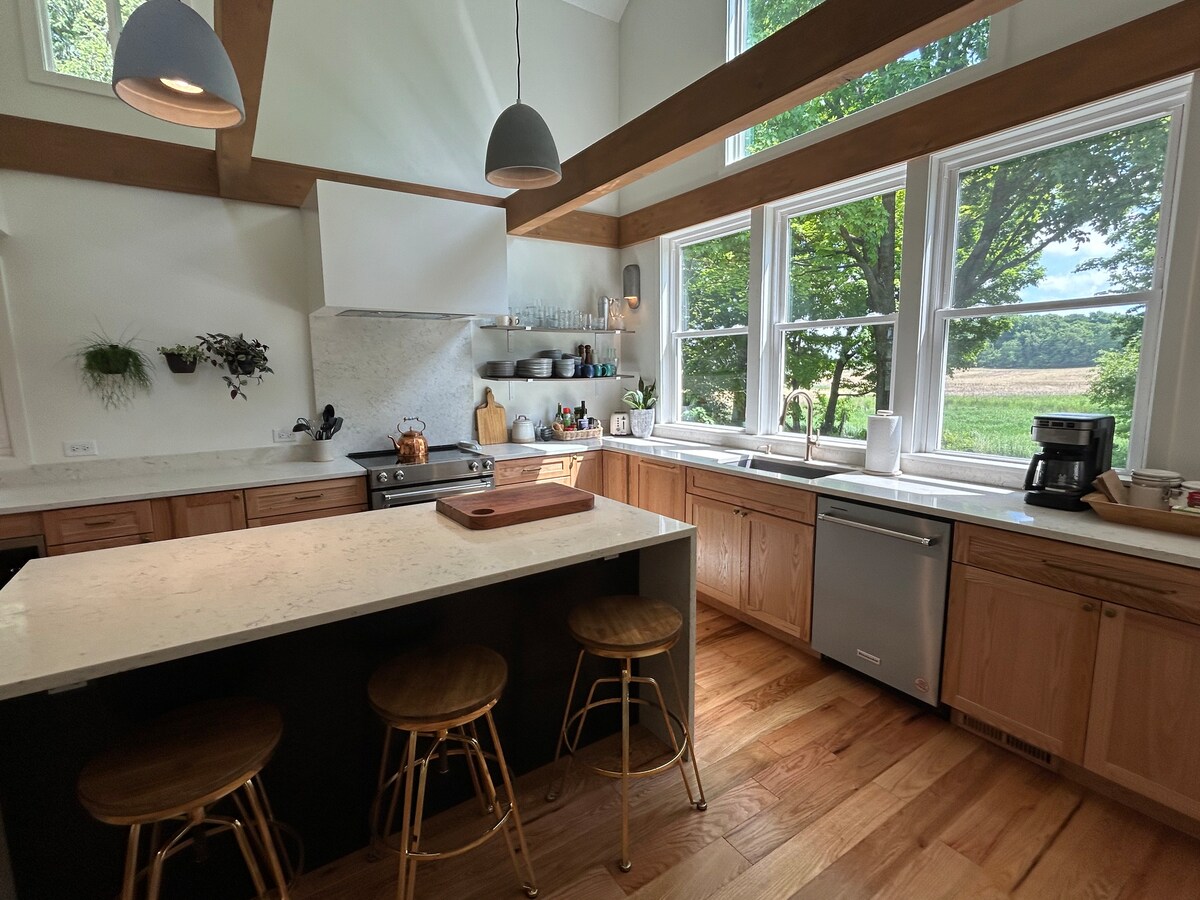The kitchen features a large island with bar seating for four, surrounded by wooden cabinetry and modern appliances. Tall windows offer views of the countryside, allowing natural light to illuminate the space. Soft pendant lights hang over the island, enhancing the kitchen's inviting design.
