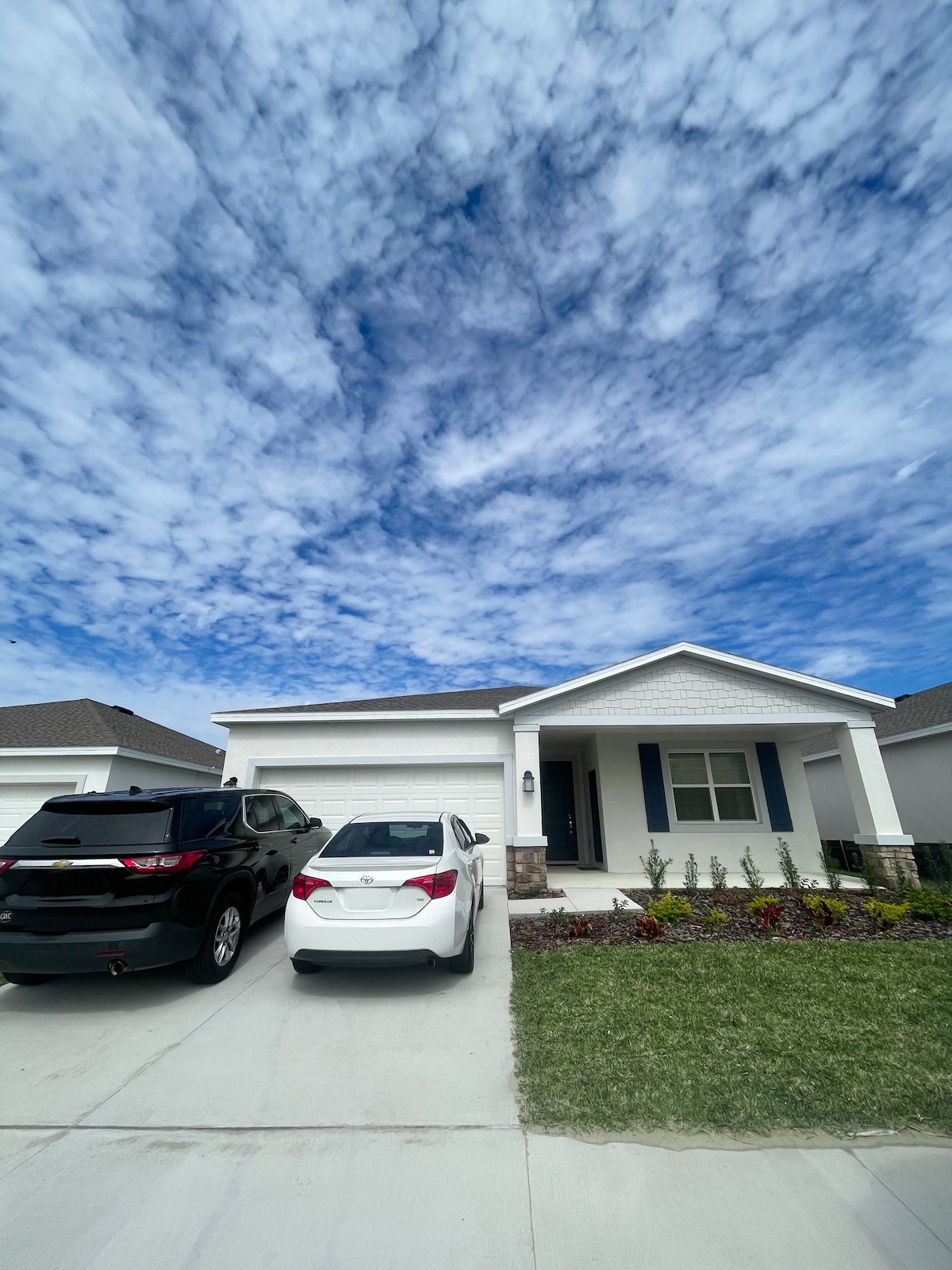 A single-story house is displayed, featuring a well-maintained exterior with a light-colored facade. The driveway accommodates two parked vehicles. Lush green grass and neatly arranged flowerbeds enhance the front landscape, while a bright blue sky with scattered clouds provides a serene backdrop.