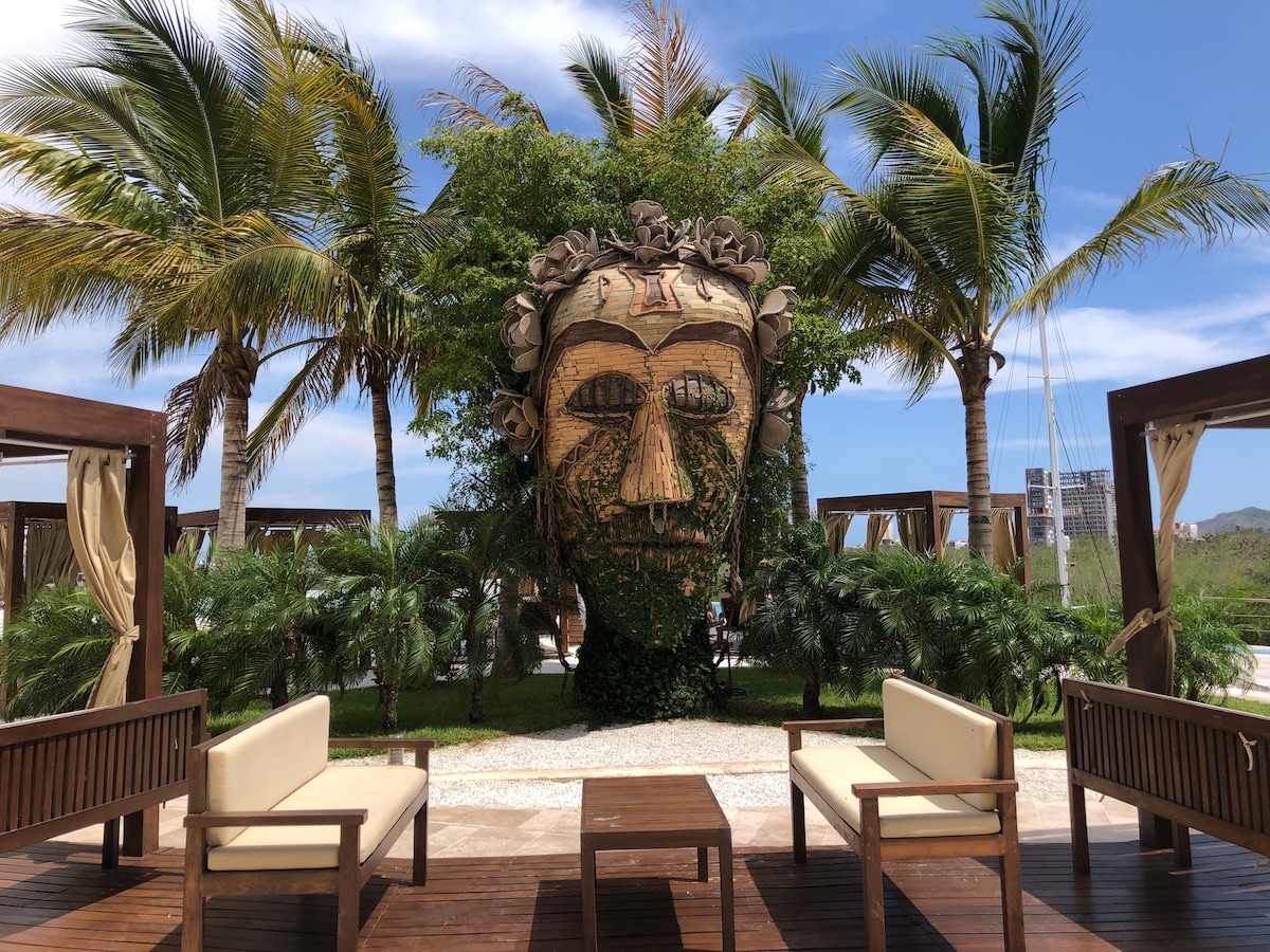 A relaxation area is presented, featuring two wooden chairs with beige cushions facing a tropical landscape. Lush palm trees surround the space, offering a serene environment. The wooden decking is complemented by a pathway leading towards a landscaped garden.