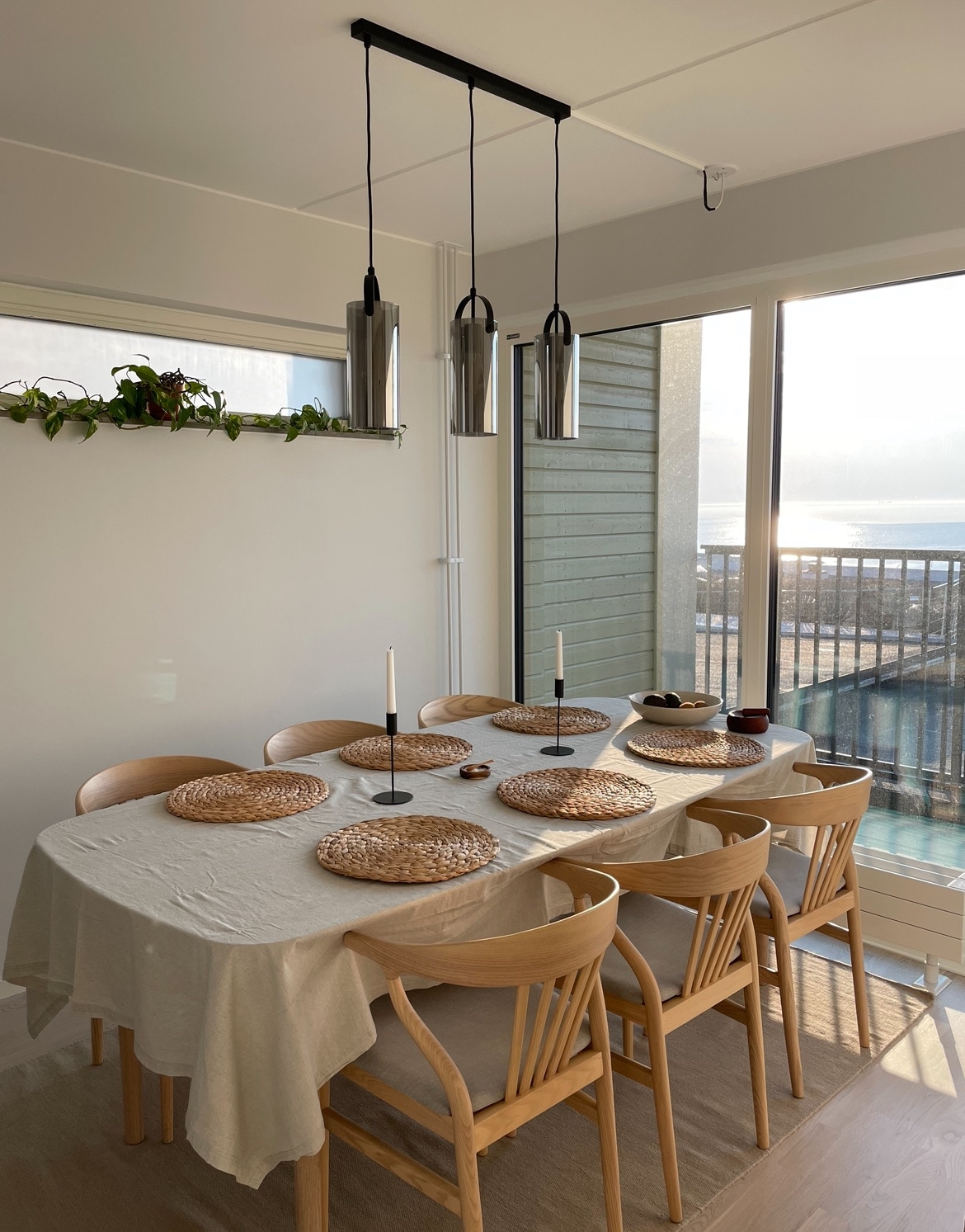 A dining area features a long table set with woven placemats and simple white dishes. Wooden chairs surround the table, while three modern pendant lights hang above. A view of the ocean can be seen through large windows, enhancing the ambiance of the space.
