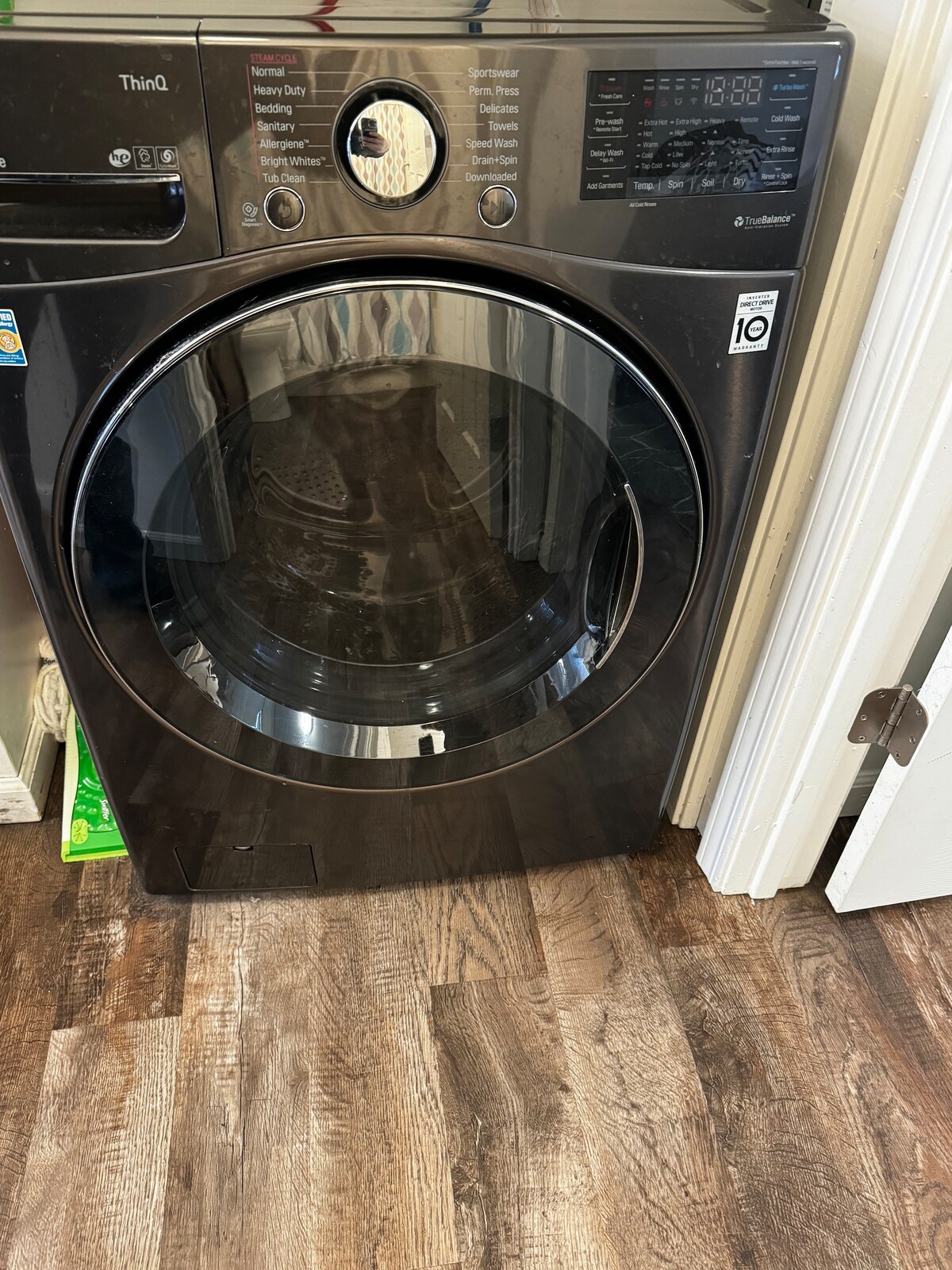 A modern front-loading washer is positioned on a wood-patterned floor. The machine features a digital display and control panel with various settings labeled. A portion of the surrounding area shows green storage bins, suggesting a utility or laundry space.