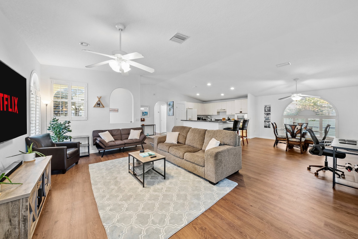 A spacious living area features a light-toned sofa and matching armchair, surrounded by wood flooring. A large area rug lies under a coffee table, while a flat-screen television displays a streaming service. The open layout connects to a dining area and a modern kitchen.