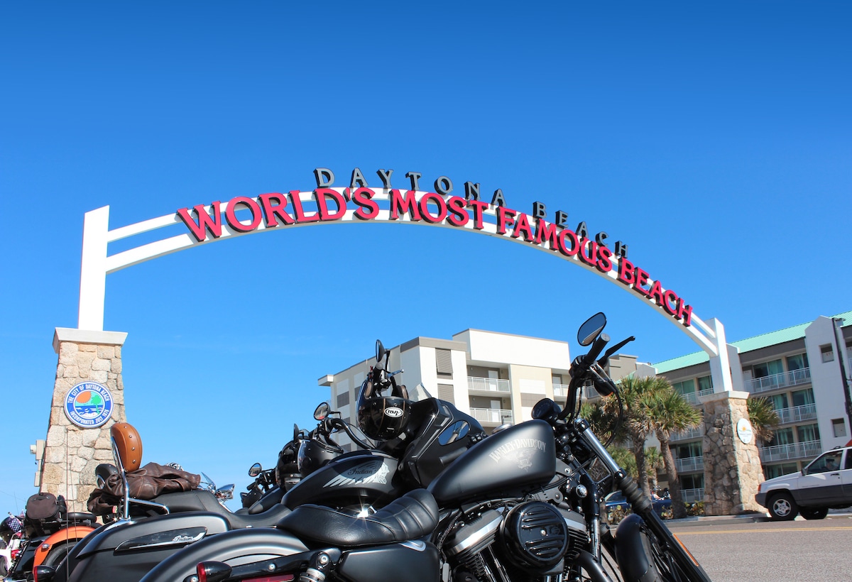 The entrance to Daytona Beach is highlighted by a prominent archway, displaying the words 'WORLD'S MOST FAMOUS BEACH.' Several parked motorcycles are gathered in the foreground, with a modern building visible in the background under a clear blue sky.